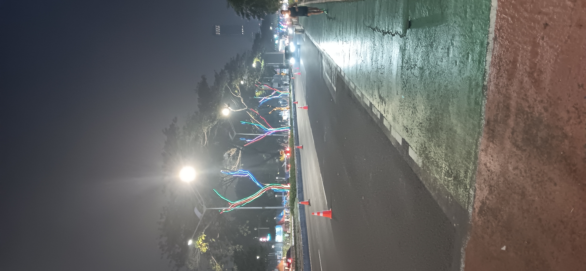 The image shows a street scene at night in South Jakarta, Indonesia. The street is lined with trees decorated with colorful LED lights, casting a vibrant glow on the surroundings. The road is wet, reflecting the lights from the streetlights and the trees. Orange traffic cones are placed along the side of the road, possibly indicating a temporary closure or hazard. In the background, a person is jogging on a green pathway next to the road, and a car is driving by with its headlights on. A tall building is visible in the distance. The sky is dark, suggesting it is late evening or night. The overall atmosphere is one of urban nightlife with decorative illumination.
