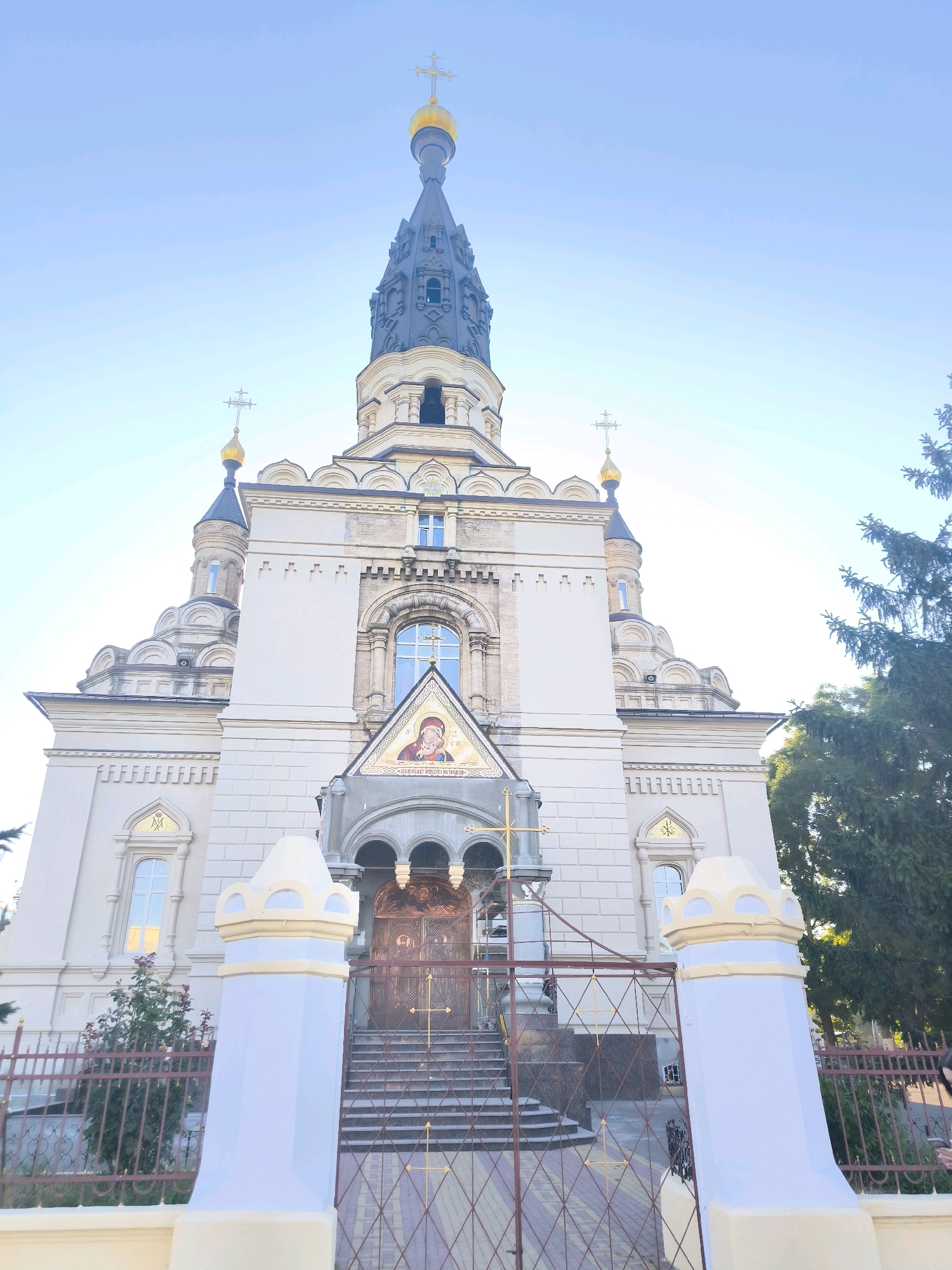 The image captures the exterior of a large, ornate church with a tall, dark spire topped with a golden cross. The building is constructed of pale stone with decorative architectural elements, including arched windows and multiple domes. A central mosaic depicting a religious figure is above the main entrance, which features ornate double doors. The church is fronted by a fence and a gateway with a golden cross motif. The sky is clear and blue. A large evergreen tree is visible to the right of the church. The church is located in the city of Mykolaiv, Ukraine.
