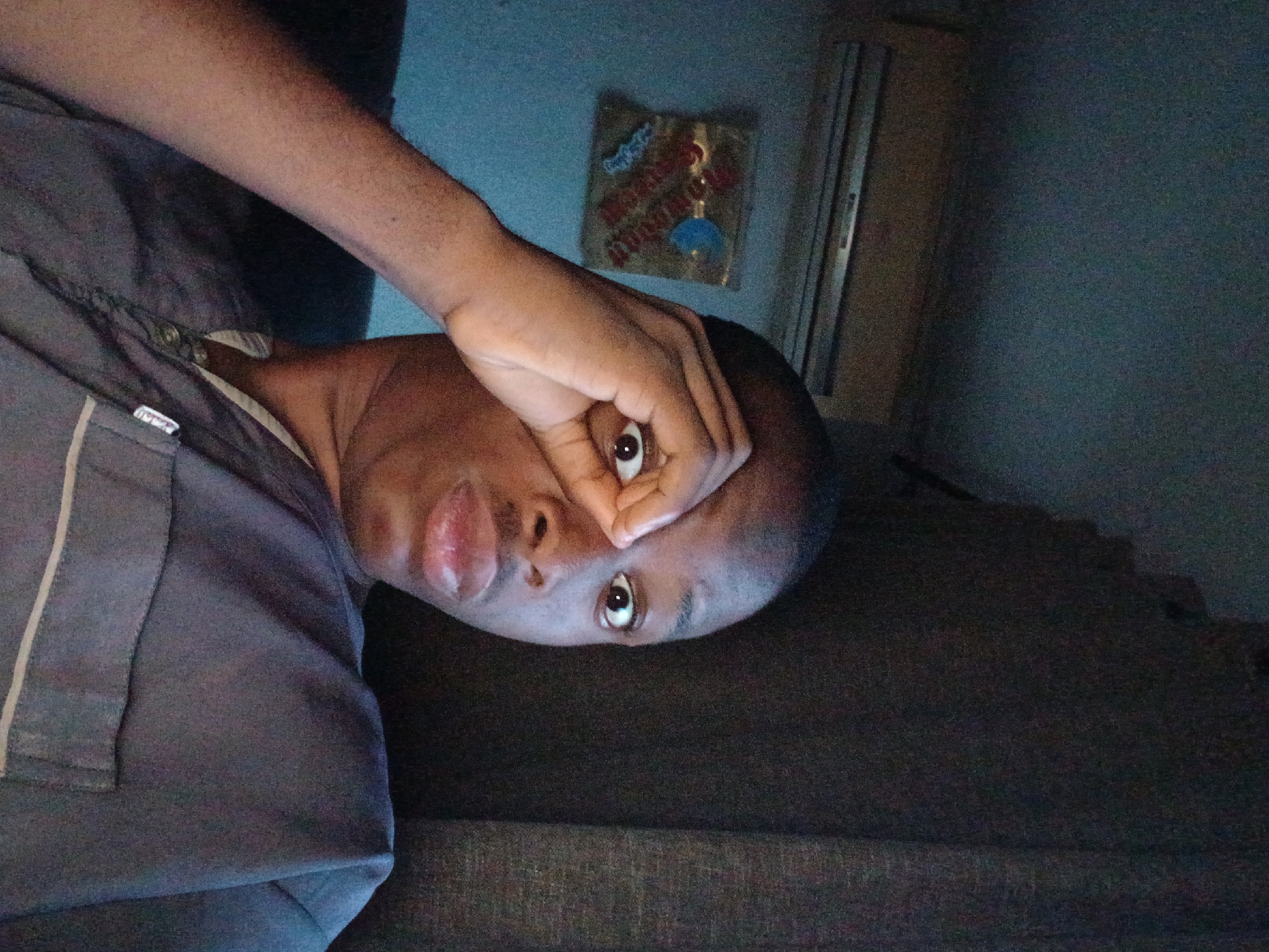 A man is holding his hand up to his eye and looking at the camera. The man is wearing a grey shirt. The background includes a wall with an object hanging on it and a sliding door. The location is likely inside a building in Lagos, Nigeria, but this cannot be determined with certainty from the image alone.
