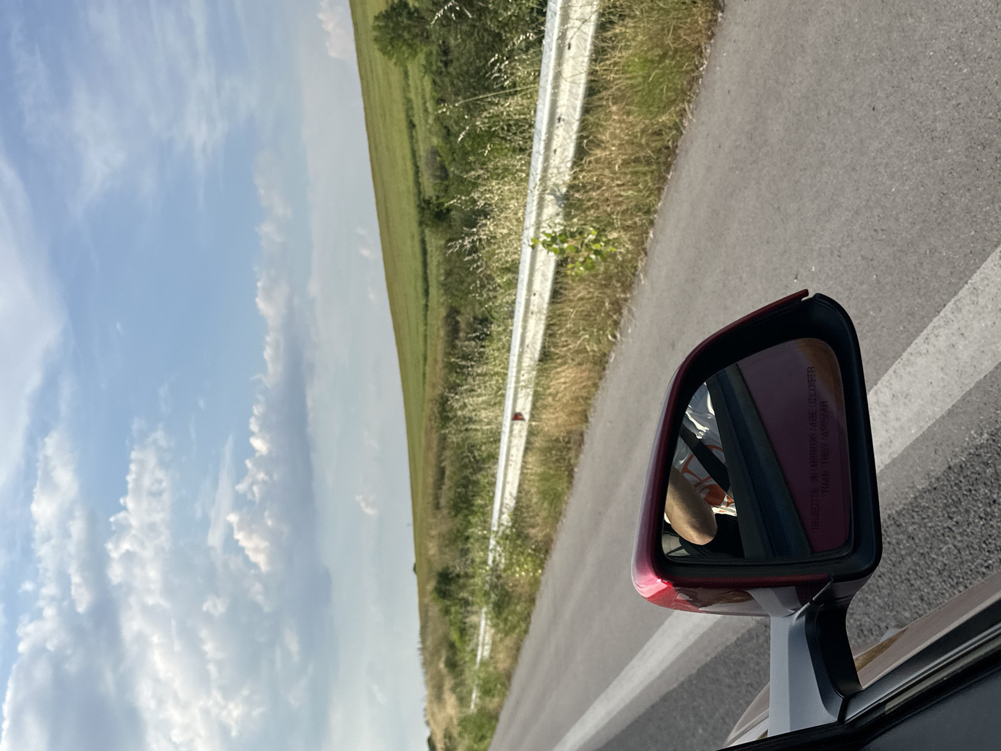 The image depicts a daylight scene observed from the front passenger side of a moving vehicle in the vicinity of Kapitan Andreevo, Bulgaria. The primary view extends along an asphalt road with a solid white line marking its right edge. To the right of the road, a white metal guardrail is present, partially obscured by an abundance of dry, yellowish grass, weeds, and green shrubbery growing roadside and behind it. Beyond the guardrail and immediate vegetation, the landscape features an expansive, upward-sloping green field or hill, indicating a rural or semi-rural environment. The distant horizon is characterized by the contours of these fields, with a few small trees visible. The sky is bright blue with scattered white cumulus clouds, suggesting fair weather.
In the foreground, occupying the lower-left quadrant, is the exterior side mirror of the vehicle. The mirror's casing is a dark red or maroon, mounted on a black base attached to the vehicle's dark-colored door frame, which is partially visible. The mirror's reflection shows the right arm and shoulder of an individual, presumably the driver, wearing a light-colored t-shirt with indistinct orange or yellow patterns. A dark seatbelt is diagonally visible across the person's shoulder and chest. The reflection also reveals part of the vehicle's interior, appearing dark red, likely a dashboard or door panel. Text is visible on the lower surface of the mirror, stating "OBJECTS IN MIRROR ARE CLOSER THAN THEY APPEAR." The texture of the asphalt road surface is visible in the lower right and foreground, appearing coarse and grey.