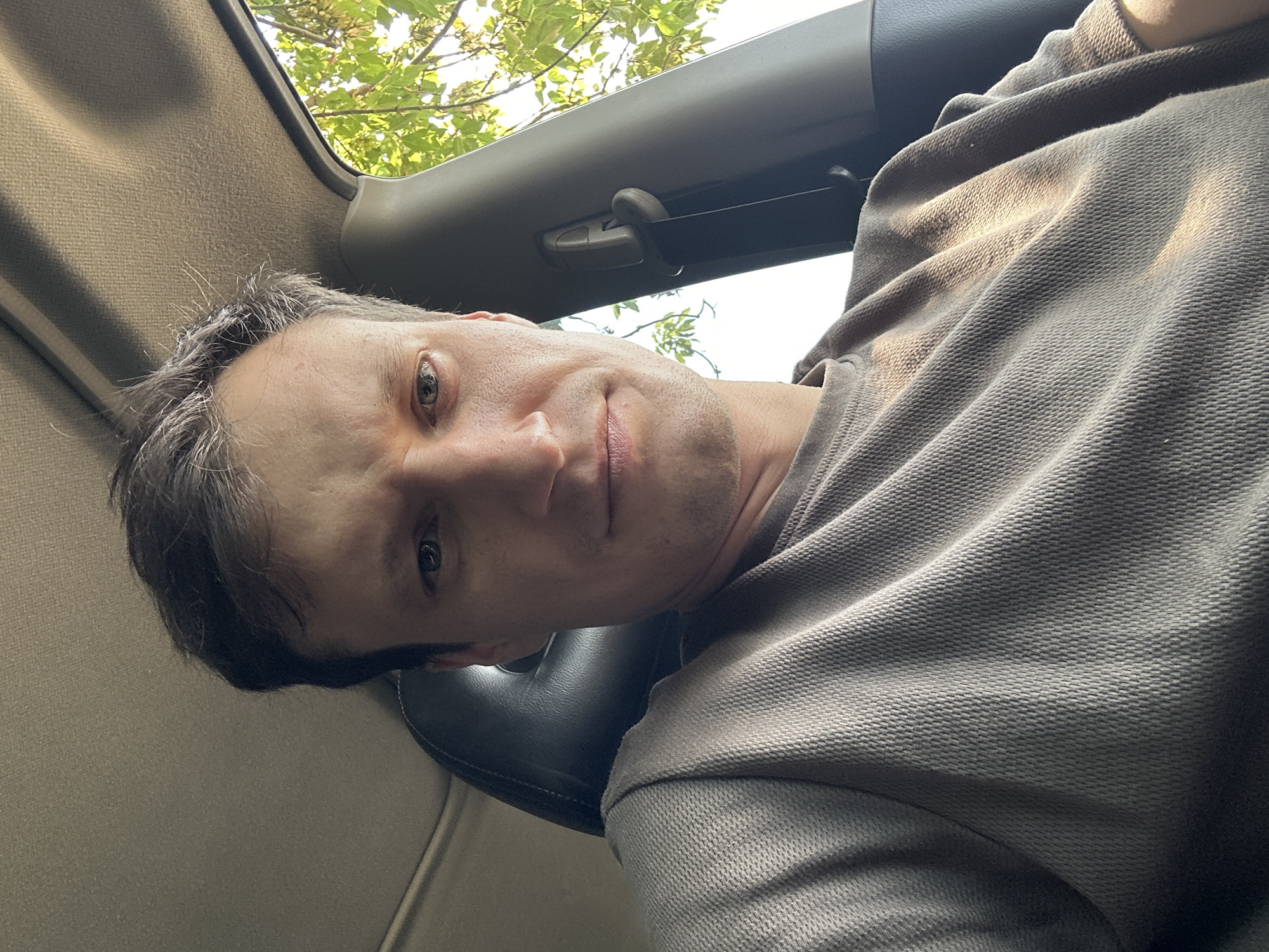 A selfie shows a man sitting in the driver's seat of a car. He has short, dark hair and is looking directly at the camera. He is wearing a light brown, textured long-sleeved shirt. The car's interior is visible, including a dark headrest and the gray headliner of the ceiling. Through the passenger window, green leaves and sunlight can be seen, suggesting an outdoor location, possibly a road passing through a wooded area or park. The image was taken in Makiv, Ukraine.