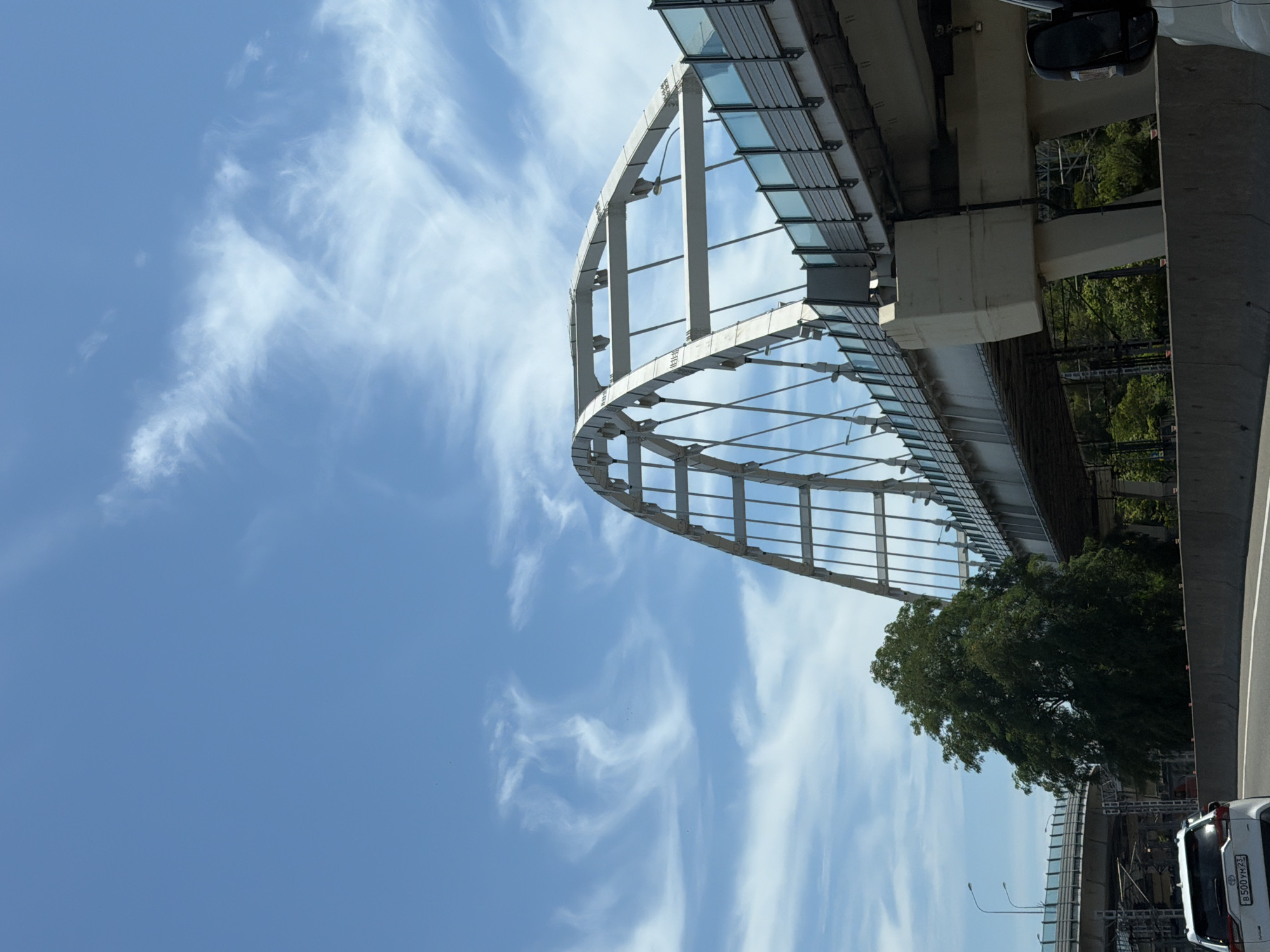 The image captures a contemporary bridge structure in Nizhnyaya Shilovka, Russia, on a clear day with wispy white clouds against a bright blue sky. The bridge features an arched superstructure made of white metal beams and supports, with modern glass barriers along its sides. Below the main bridge, another elevated road or structure is visible, also with concrete supports and a similar roadway. A large, dense green tree stands prominently near the base of the main bridge. In the foreground, the rear of a white SUV with a visible license plate (B 500 YM 23) is positioned on a multi-lane road. The road is divided by concrete barriers, and a portion of another vehicle, possibly a van or truck, is visible in the lower right corner. The overall scene depicts urban infrastructure in a well-lit environment.