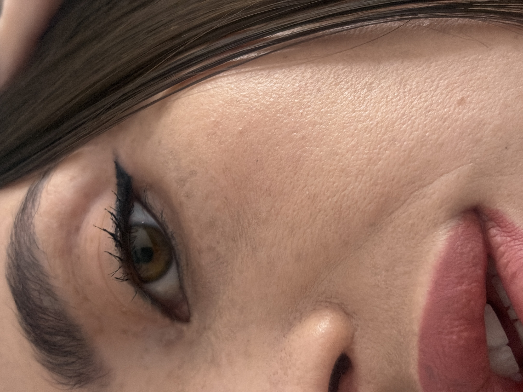 An extreme close-up portrait features the right side of a person's face. The dominant elements are the right eye, a portion of the nose, and the upper lip with a visible segment of the lower lip. The individual's right eye displays a brown iris, dark eyelashes enhanced with black mascara, and a sharp black winged eyeliner application. The eyebrow above is dark brown and well-defined. The skin, a light to medium tone, shows visible pores and fine lines. The bridge of the nose and the right nostril are discernible. The lips are coated with a matte, pinkish-rose lipstick, and the mouth is slightly agape, revealing white upper and lower teeth. Dark brown hair frames the right side of the face. The background is a uniformly light, out-of-focus area, offering no specific environmental details. There are no visual indicators within the image to identify its location as Saint Petersburg, Russia, or any other specific city.