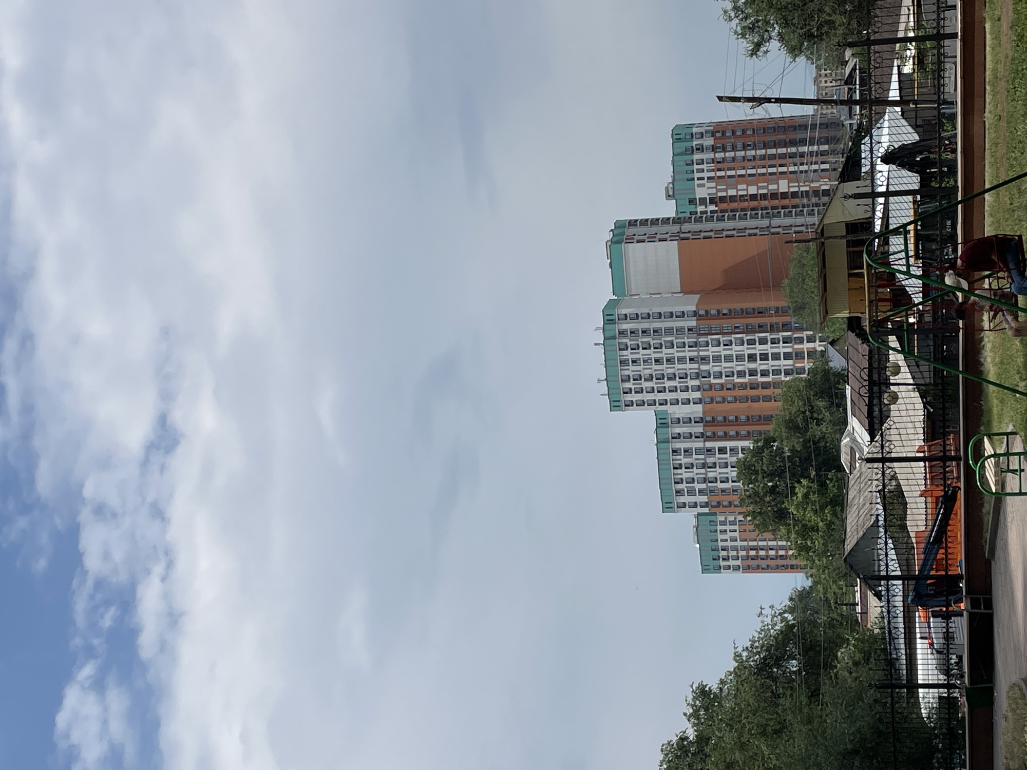 The image shows a residential neighborhood in Almaty, Kazakhstan, with tall apartment buildings in the background and a playground in the foreground. The apartment buildings are modern, with white, orange, and teal facades. They are arranged in a cluster, with trees and other buildings in between. In the foreground, there is a playground with a swing set. Two children are on the swing set, and there is a person sitting on the grass nearby. The sky is cloudy, and the weather is overcast. The overall impression is of a typical urban residential area.