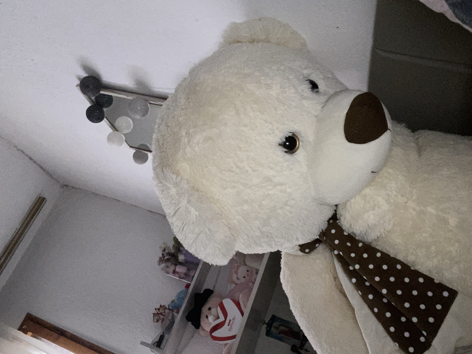 The image shows a close-up of a large, white teddy bear with a brown nose and a brown and white polka-dot scarf. The teddy bear appears to be positioned at an angle, looking down. In the background, there's a glimpse of a white wall, a shelf with various stuffed animals, and a modern light fixture with spherical bulbs. Due to the limited context and lack of identifying details, I am unable to determine the specific location in Blagoevgrad, Bulgaria.