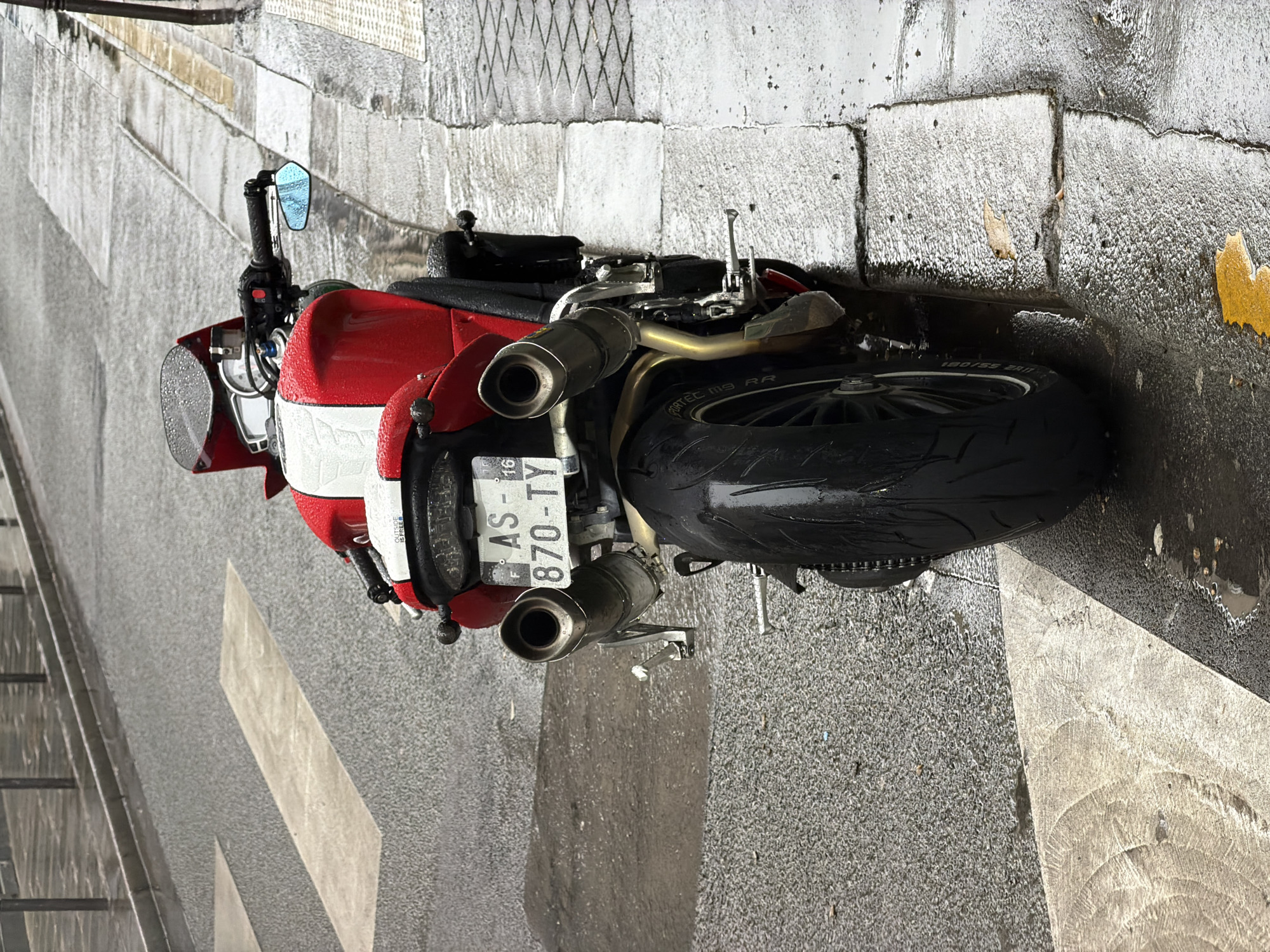 The image shows a wet street scene in Paris, France, with a parked red and white motorcycle as the central subject. The motorcycle, displaying license plate "AS-870-TY," is parked on the edge of a crosswalk, adjacent to a sidewalk made of rectangular stone blocks. The street and sidewalk are glistening with moisture, indicating recent or ongoing rain. The background features a wall constructed of similar stone blocks. There are no people visible in the frame.