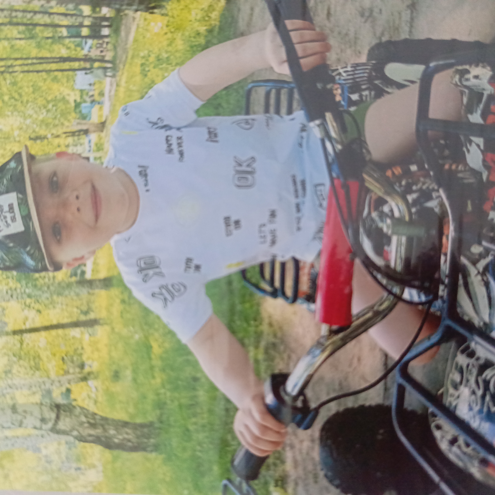 A young boy is sitting on a quad bike in an outdoor setting. He's wearing a white t-shirt with graphic prints and a baseball cap. The background appears to be a grassy area with trees. The location is Unknown, Unknown.