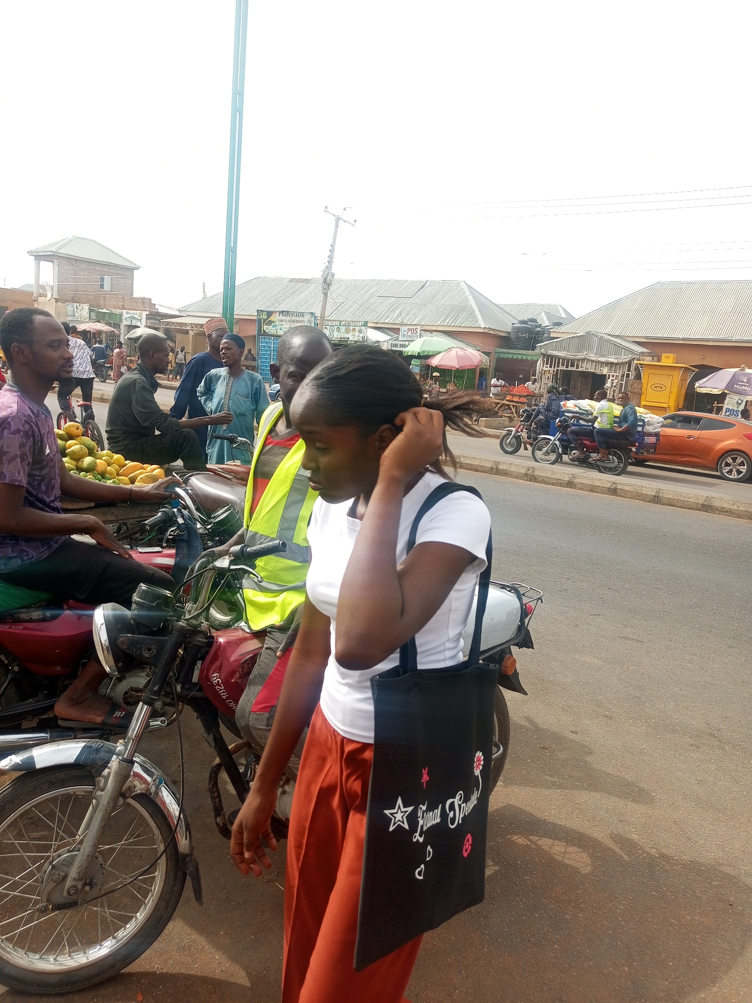 This image captures a vibrant street scene in Tempure, Nigeria, depicting daily life and commerce under what appears to be a bright, sunny sky. In the foreground, a young woman, seen from the side, walks or pauses. She wears a white t-shirt, orange pants, and carries a black tote bag with white text "Eternal Spirit" along with star, flower, and heart motifs. Her hair is pulled back.
Behind her, the scene is bustling with activity. A man sits on a red motorcycle, positioned next to a large display of yellow fruits, resembling mangoes, indicating a fruit vendor. Another man, wearing a bright yellow safety vest, is also seated on a motorcycle nearby. Several other men are visible in the mid-ground and background, some conversing, others on motorcycles, and one in blue traditional attire.
The setting is characterized by a roadside market or commercial area with various small businesses. Buildings with tin roofs line the street, and market umbrellas provide shade for some stalls. Overhead, power lines are visible. An orange car can be seen in the distance on the road.
Notable visible text includes "PROVISION," "SHINING INGREDIENTS," "CHINONSO RESTAURANT," "POS" (Point of Sale), "CAKE HOU" (likely "Cake House"), and the branding "MTN" and "MoMo," indicating mobile communication and money services. A red motorcycle has the numbers "10 18239" visible, possibly a registration number. The image conveys an active, everyday atmosphere in an urban or semi-urban Nigerian setting.