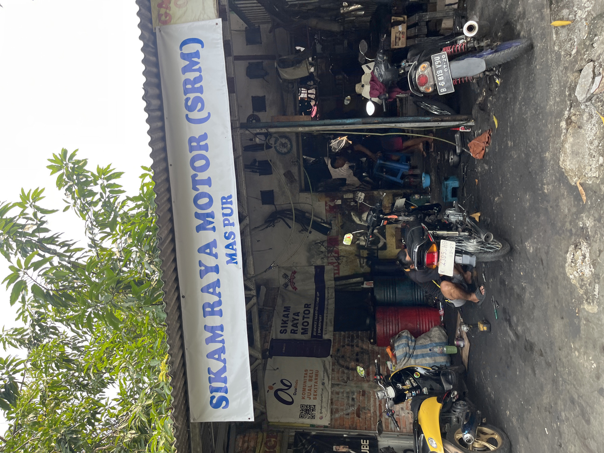 An exterior, medium shot shows a motorcycle repair shop in Tangerang, Indonesia. The shop features a corrugated metal roof and brick walls. A banner across the front reads "SIKAM RAYA MOTOR (SRM) MAS PUR" in blue lettering. Below this, another banner displays the logo for "DuaRoda" with text in Indonesian, indicating services like sales, purchases, and exchanges. Further to the right, a banner for "SIKAM RAYA MOTOR" lists services such as "SERVICE | GANTI OLI | TUNE UP | SPAREPART" and an address in Tangerang.
Inside the shop, motorcycles are being repaired. One yellow and black scooter is parked on the left. In the center, a man is kneeling and working on a motorcycle. Another motorcycle, black with red accents, is parked on the right side of the frame. Various tools, barrels, and spare parts are visible throughout the workshop, contributing to a cluttered yet functional atmosphere. The ground is dusty and appears to be a workshop floor. Lush green trees overhang the roof of the shop.
