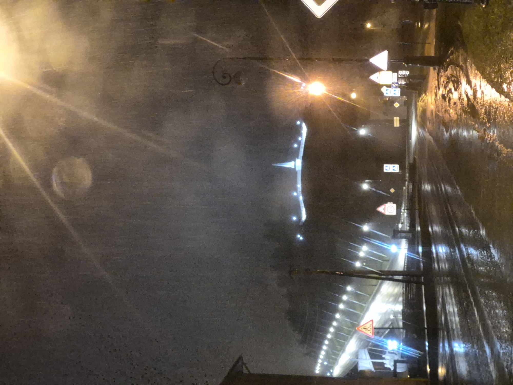 The image depicts a rainy night scene in Tobolsk, Russia. The street is wet and reflecting the lights, creating a blurred effect. The visible elements include streetlights, road signs, and an illuminated bridge in the distance. The image conveys a sense of urban landscape under adverse weather conditions.