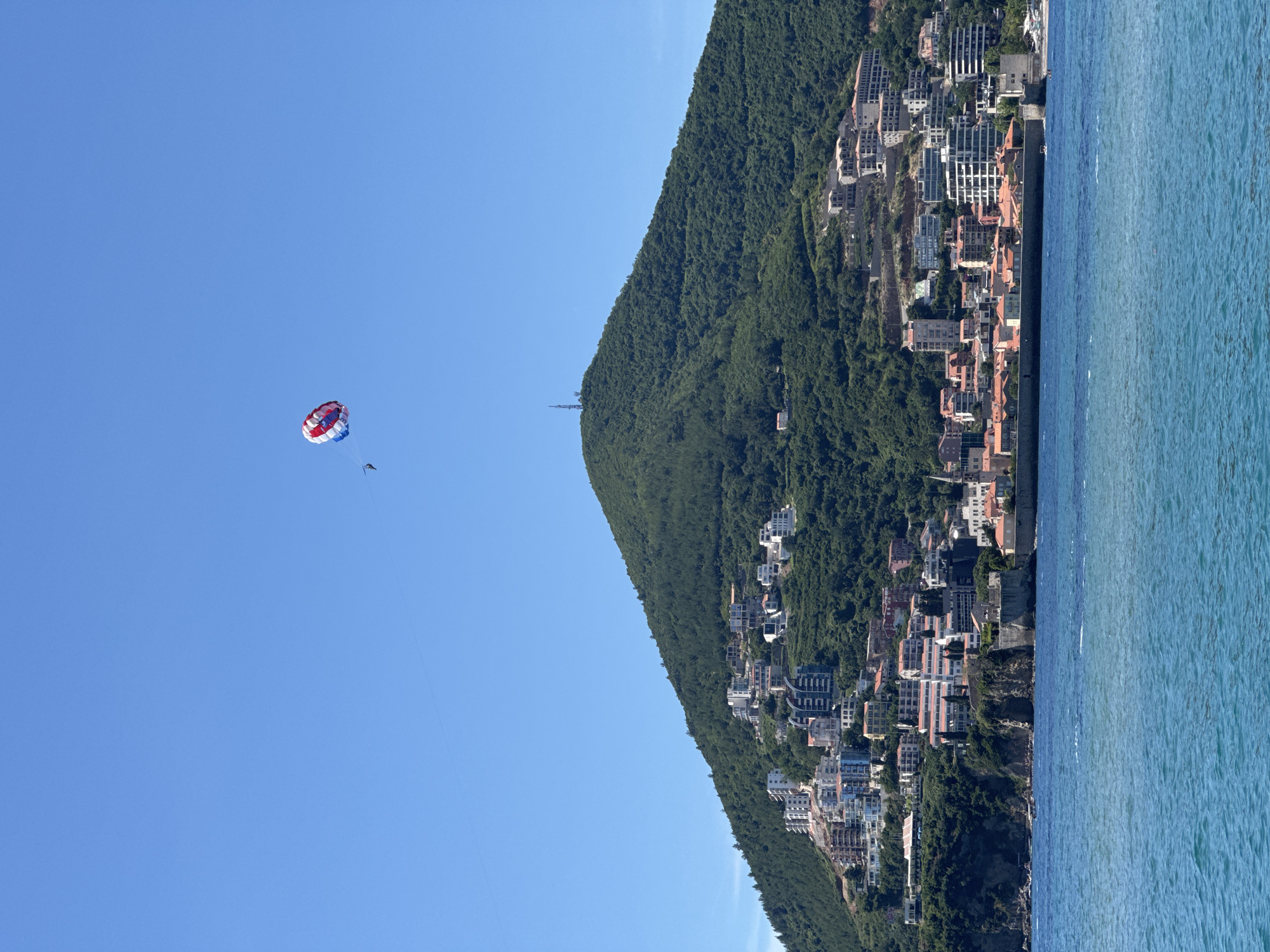 The image captures a vibrant coastal scene in Budva, Montenegro, under a bright, clear blue sky. High above the water, a person is parasailing, their parachute a striking combination of red, white, and blue. A thin tow rope extends from the parasailer.
The landscape features a large, verdant hill or mountain, densely covered in trees, sloping down towards the sea. Nestled into this hillside is a sprawling town, characterized by numerous buildings of varying sizes and styles, including residential structures and possibly hotels, many featuring light-colored facades and some with orange-ttiled roofs. A tall, slender antenna or communication tower is visible on the peak of the hill, and a smaller structure, possibly a cable car station or gondola, can be seen on the slope.
To the right, the calm, deep blue water of the sea stretches out, showing subtle ripples on its surface. The clear sky and bright illumination indicate that it is daytime, likely morning or early afternoon, under sunny and pleasant weather conditions, ideal for outdoor activities such as parasailing. The scene conveys a lively resort atmosphere. There is no visible text in the image.
