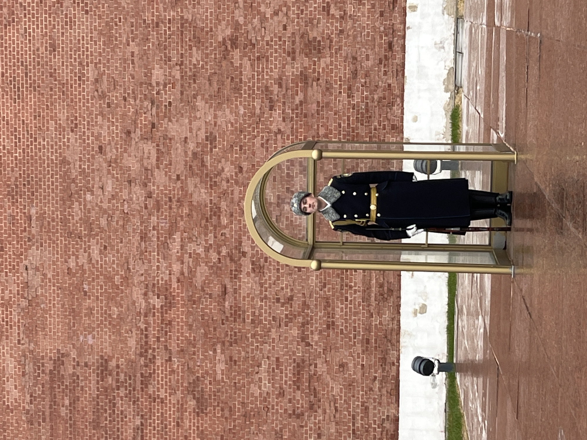 A color photograph shows a soldier standing guard at a station in front of a red brick wall. The soldier, wearing a dark blue uniform with gold buttons and a fur hat, stands at attention within a gold-framed glass enclosure. He holds a rifle in his hands. The ground around the enclosure is paved with reddish-brown stone. The background features a tall brick wall that extends into the distance. The location is unknown, but the scene is most likely Moscow.