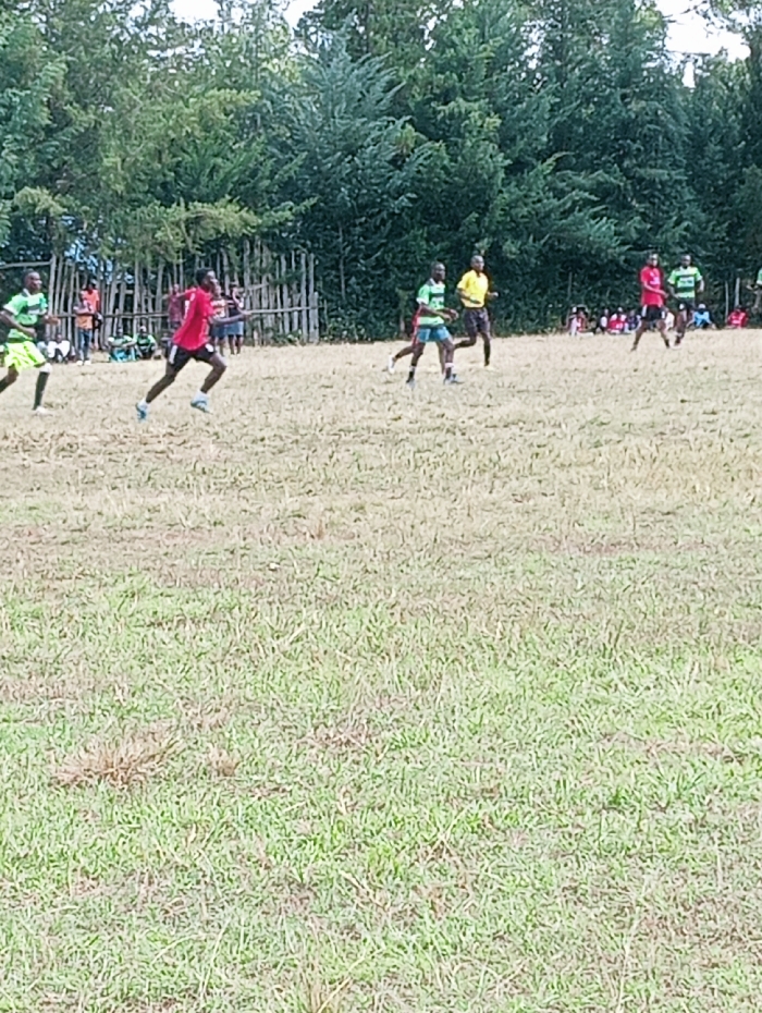 An outdoor scene in Lwandeti ward, Kenya, depicts a football match in progress on a dry, partially grassy field. The foreground consists of sparse green and brown grass. In the midground, several male individuals are actively participating: one player in a red jersey and black shorts runs from left to right, left of center; another player in a green and light blue striped jersey and dark shorts runs from right to left, right of center. Behind him, a player in a yellow jersey and dark shorts moves in the same direction. Other players in green and red jerseys are visible further back on the field. In the background, a low, rustic wooden fence separates the playing area from a small group of spectators seated or standing on the left. A dense line of various trees forms the farthest background. The lighting suggests daytime conditions.