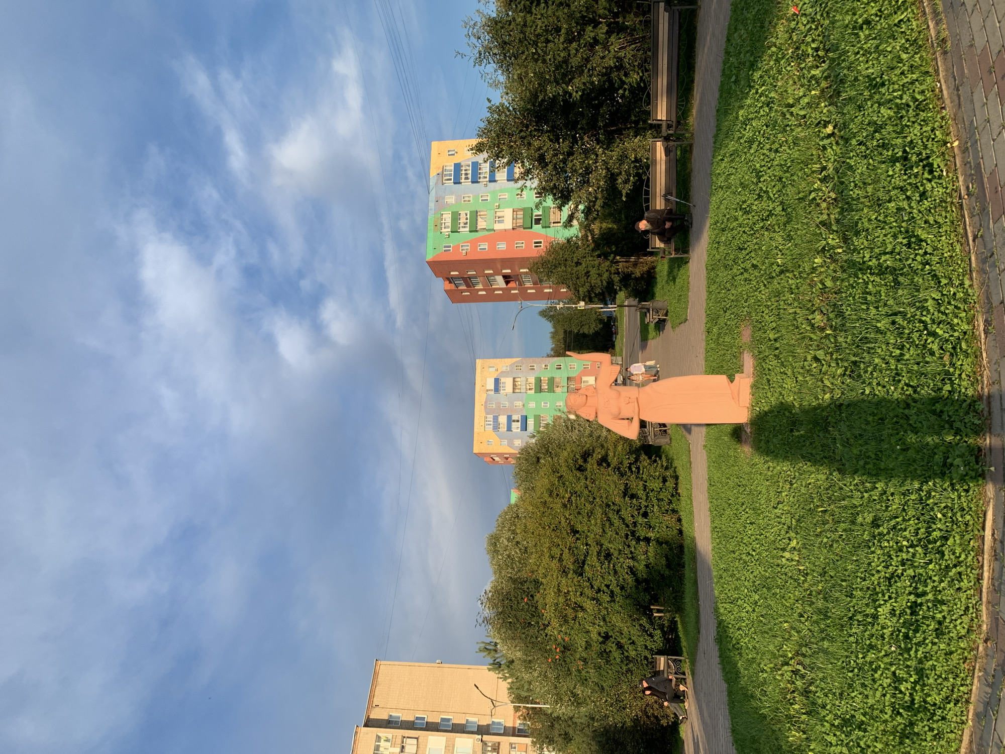 The image depicts a park scene with a statue of a woman in the foreground. The statue is light orange and casts a long shadow on the green grass. In the background, several colorful apartment buildings are visible, along with trees and benches. Some people are seated on the benches, and others are walking on a paved path. The sky is partly cloudy, with a mix of blue sky and grey clouds. The location is identified as Prokopyevsk, Russia.
