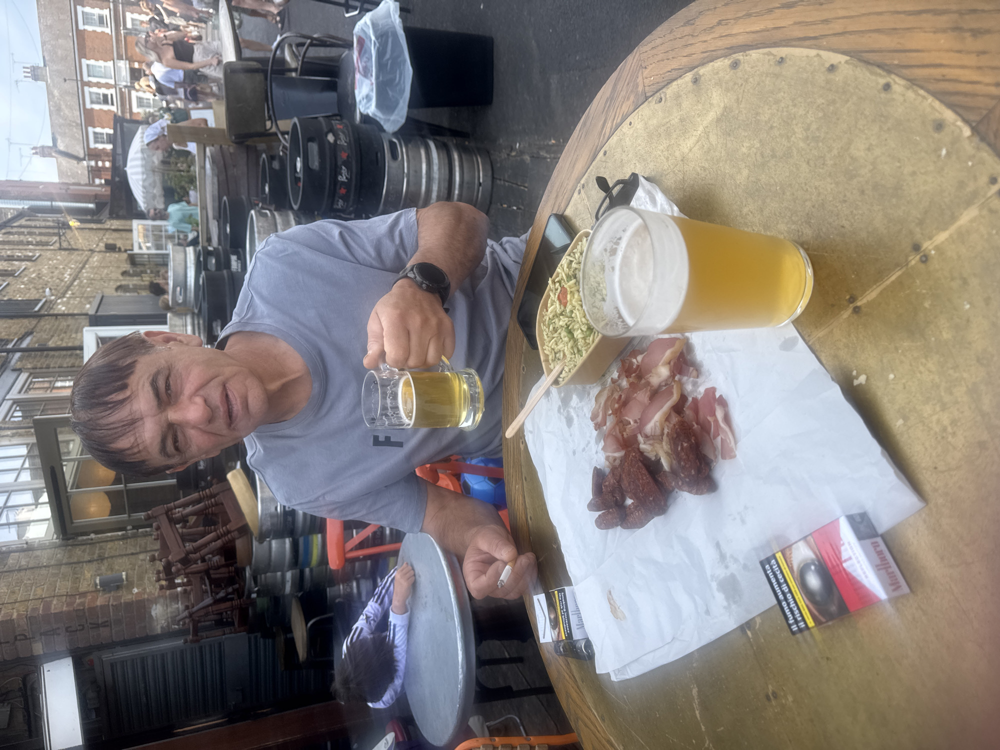 A man is sitting at an outdoor table with two glasses of beer, a plate of cured meats and what appears to be a noodle salad. In front of him is a pack of Marlboro cigarettes and a lit cigarette. He is wearing a grey t-shirt and a watch on his left wrist. In the background, there are stacks of kegs, chairs, and people walking around. The setting appears to be a pub or beer garden in London, United Kingdom. The man is looking directly at the camera, holding one glass of beer in his right hand and a cigarette in his left. To the left of the table, a child is lying face down on another table.