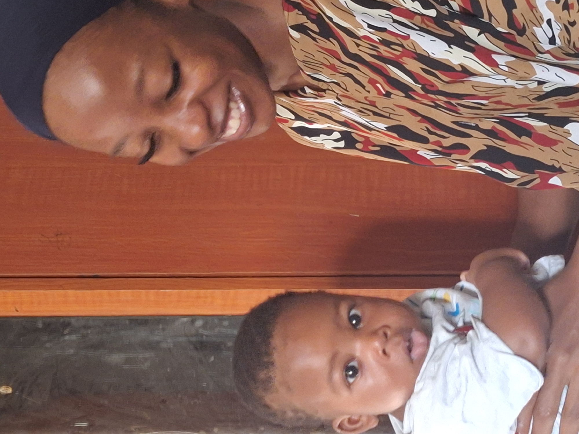 A woman with dark skin and a black head covering smiles with her eyes closed. She wears a patterned top in shades of brown, black, white, and red. She is holding a baby, who has dark skin and hair. The baby is looking directly at the camera, sticking its tongue out slightly. The baby wears a white shirt. The background appears to be a wooden wall and a dark surface, possibly a countertop or table. Yelwa, Nigeria is not discernible in the image.
