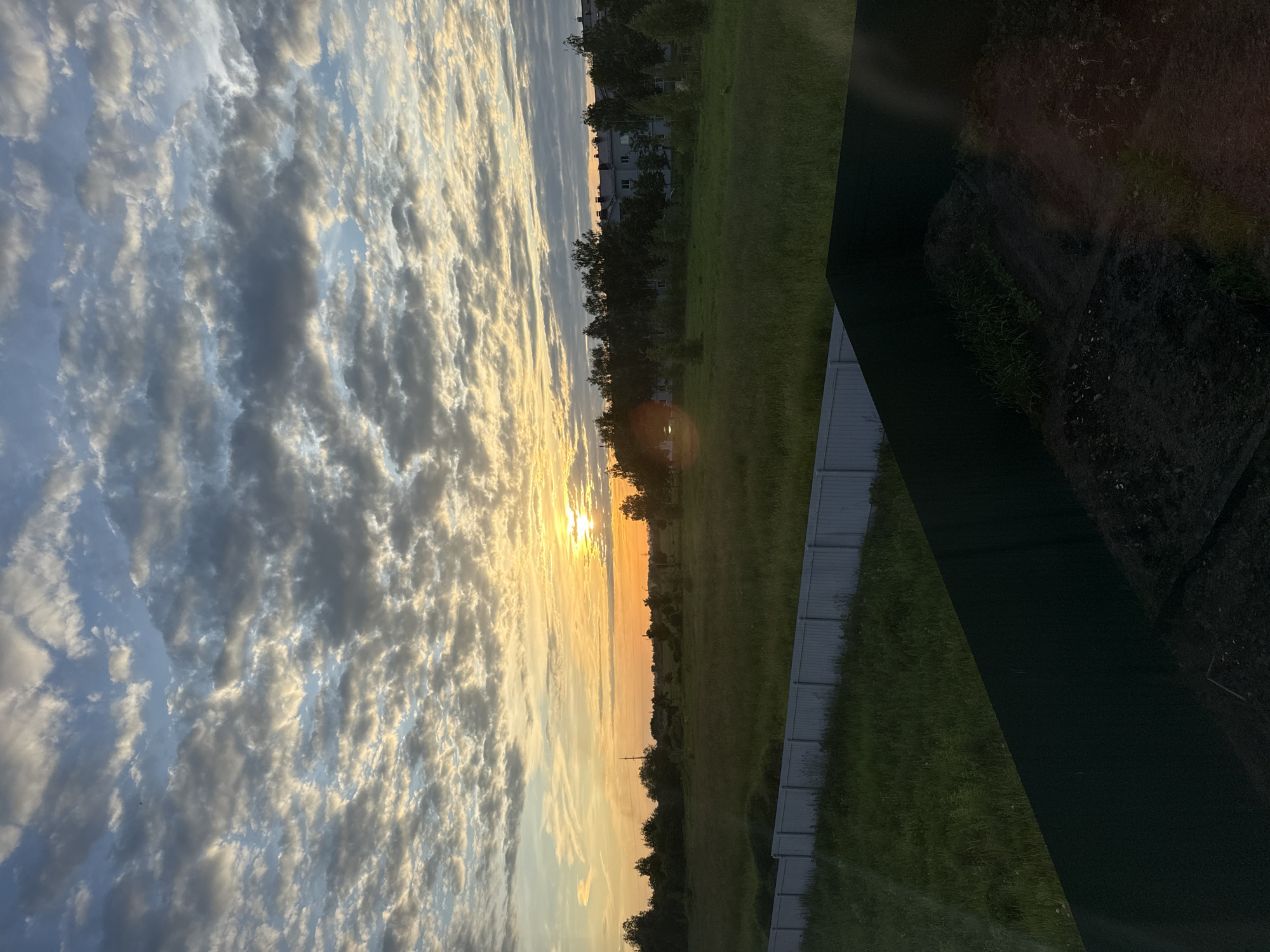 This landscape photograph captures a vibrant sunset or sunrise over a semi-rural scene in Murom, Russia. The upper portion of the image is dominated by a dynamic sky filled with altocumulus or stratocumulus clouds. The left side of the sky is a clear light blue, gradually transitioning towards the right where the low-hanging sun bathes the clouds in warm hues of orange, yellow, and golden light. A bright sunburst and lens flare are visible near the horizon, partially obscured by a distant line of dark green trees.
Below the horizon, a strip of lush green grassland stretches across the mid-ground. Faintly visible behind the treeline are a few low-rise buildings, suggesting a residential or suburban area in the distance. In the lower part of the frame, a light-colored fence, possibly made of corrugated metal or similar material, runs horizontally. More green grass is present in front of and behind this fence. A dark, angled shape occupies the bottom right corner, likely a window frame or part of the interior from which the photograph was taken. The image conveys a sense of serene natural beauty during the golden hour. No people or visible text are present.