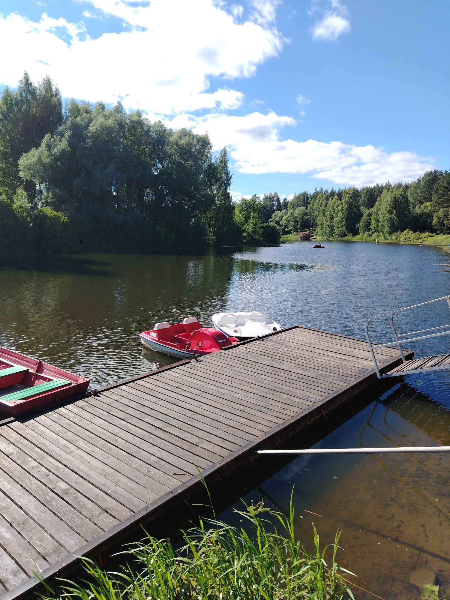 The image shows a wooden dock on a lake, with several pedal boats moored to it. Two red and white pedal boats are clearly visible, and part of a white pedal boat is also in view. The dock is made of weathered wooden planks and has a metal handrail and ramp leading to the water. The lake is surrounded by lush green trees, and the sky is blue with scattered white clouds. In the distance, several other pedal boats and what appears to be a small structure are visible on the water. The scene suggests a recreational area, likely within or near the city of Stroitel', Russia, given the context. The water is dark and reflects the trees and sky. Green reeds and aquatic plants are growing along the edge of the dock.