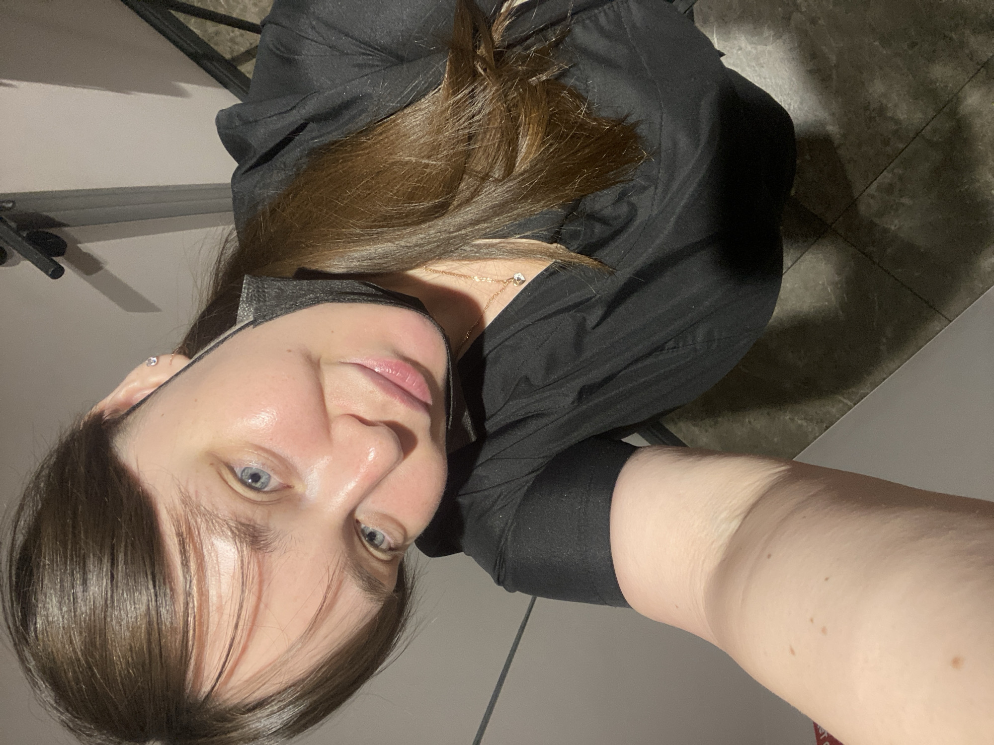 A selfie-style image depicts a woman with light skin, blue eyes, and brown hair. She is wearing a black shirt and a dark face mask partially covering her face. A delicate gold necklace is visible. The background consists of a wall on one side and a tiled floor on the other. The location cannot be determined from the image, and no connection to Votkinsk, Russia, can be established.