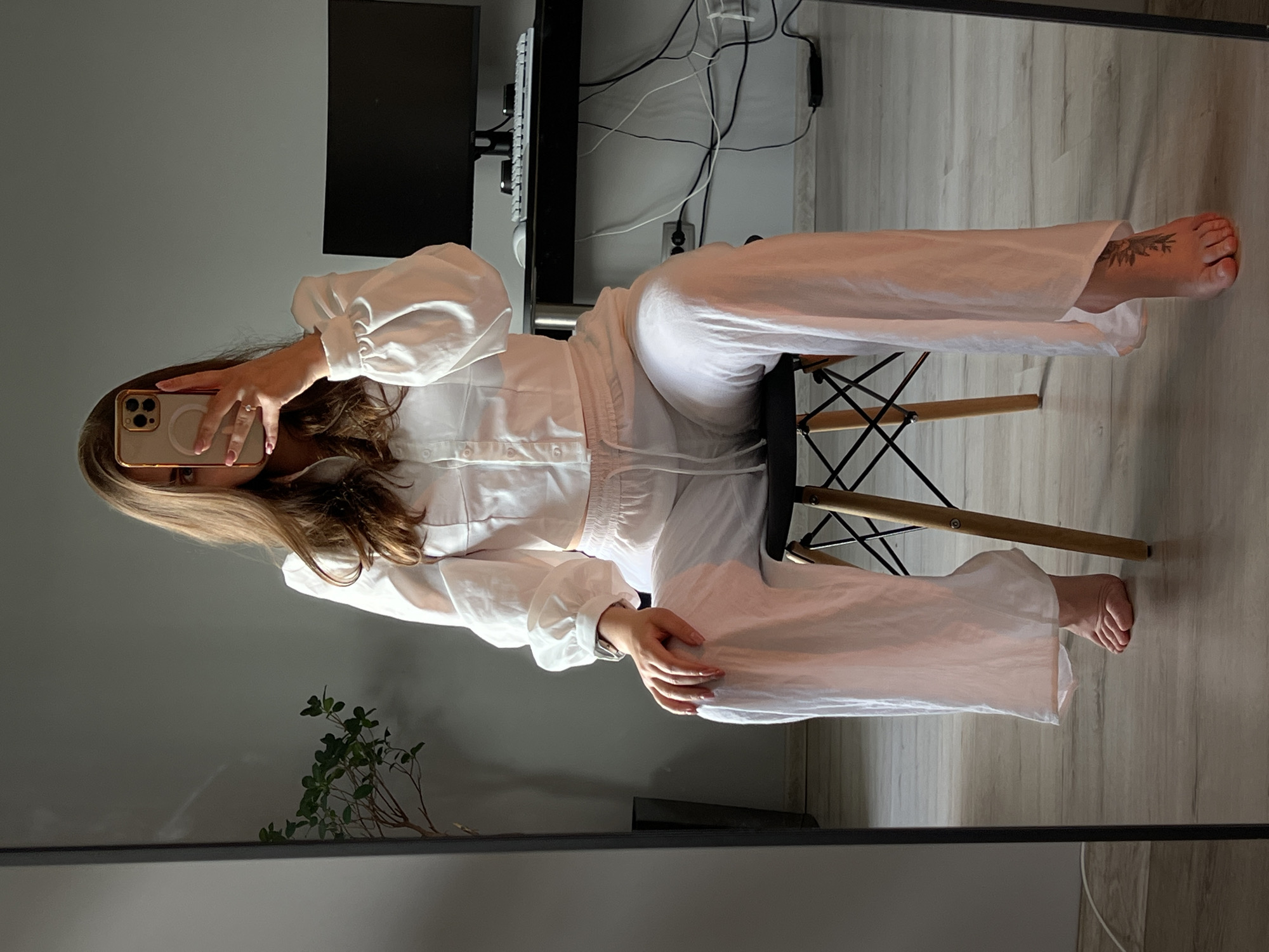A female individual is depicted taking a mirror selfie in an indoor setting in Ternopil, Ukraine. The individual, appearing to be an adult with long, light brown hair, is partially obscured by a gold-framed smartphone held in her right hand, which displays a circular MagSafe accessory. She is wearing a white, collared, long-sleeved button-up shirt with voluminous sleeves and white drawstring wide-leg pants. Her fingernails are painted light pink. She is seated barefoot on a black-seated chair with wooden legs. A tattoo, resembling a botanical or floral design, is visible on the dorsal aspect of her right foot.
The background, reflected in the mirror, shows a room with light wooden flooring and grey walls. To the viewer's left, a potted plant with green foliage is visible in a dark planter. To the viewer's right, a black desk with a black computer monitor, a white keyboard, and a white mouse is present. Beneath the desk, a cluster of black and white electrical cables is connected to a white wall outlet. A small black electronic device rests on the floor near the cables. The room is illuminated by natural light, casting shadows.