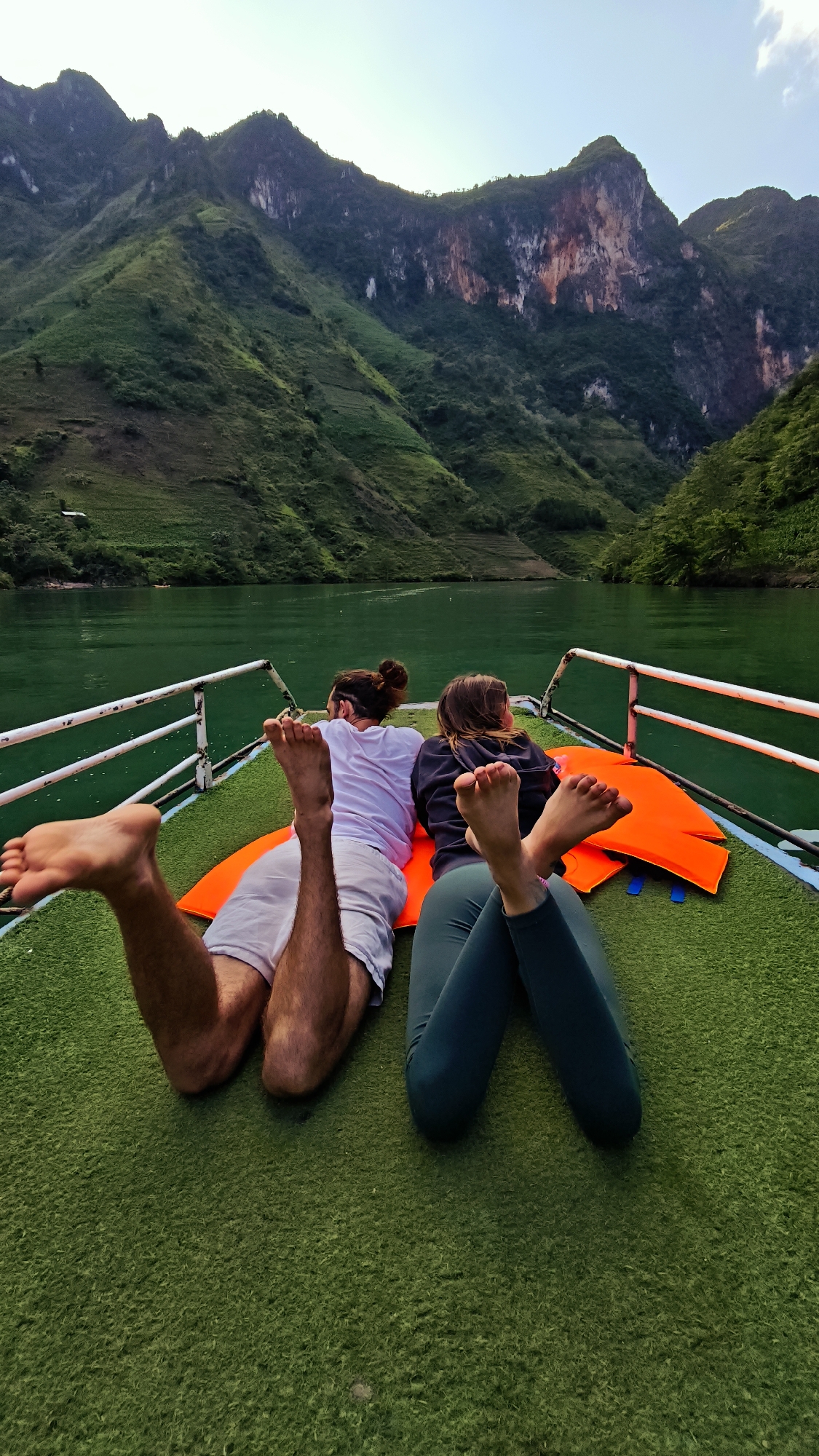 Two people, a male and a female, are lying on their stomachs on the deck of a boat. An orange life vest is underneath them. The male is wearing white shorts and a white shirt, while the female is wearing dark green leggings and a dark hooded sweatshirt. The boat deck is covered in green turf. Behind the boat are mountains with lush green vegetation. The water is a murky green color. The location is in the city of Đồng Văn Commune, Vietnam.