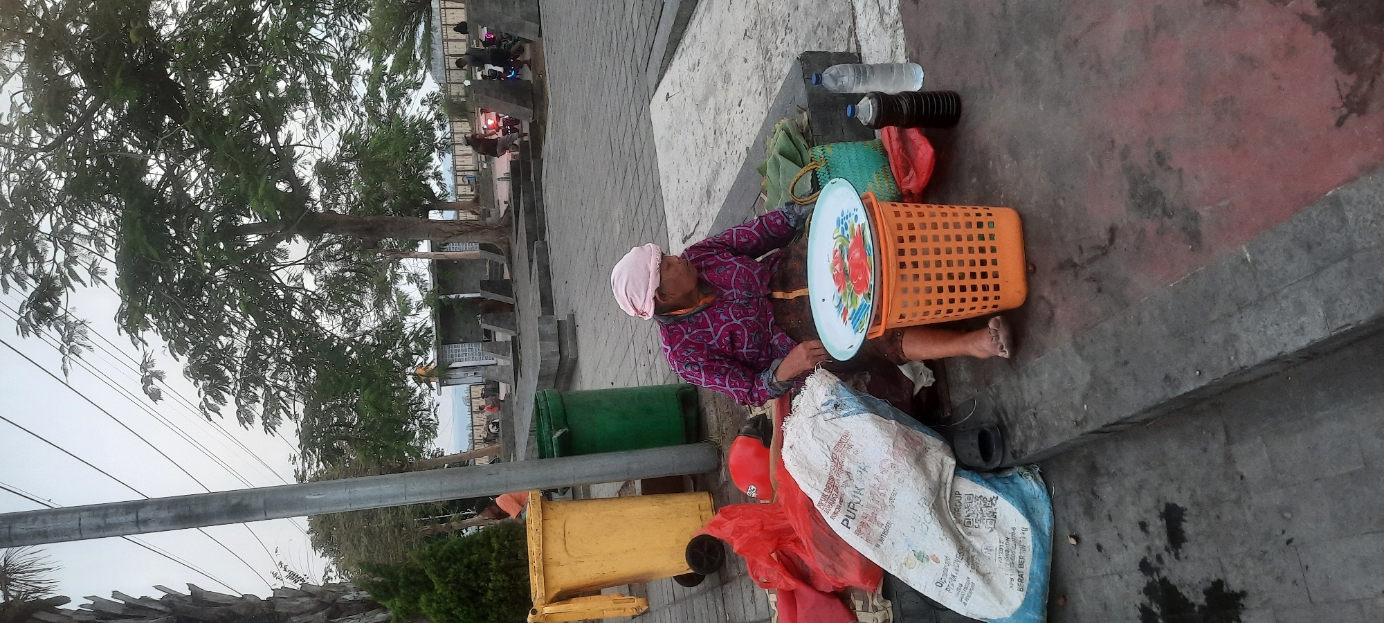 In Kanigoro, Indonesia, a woman wearing a pink headscarf and a patterned pink and purple shirt sits on a concrete ledge. In front of her, an orange plastic basket holds a decorative metal tray with floral patterns. To her right, there are two bottles: one clear and one dark, likely containing liquids, and a green woven bag. A large white sack, possibly containing fertilizer, lies to her left, next to a red plastic bag and an orange helmet. Nearby, a yellow trash can and a green trash can are visible. In the background, several people are present, some near motorcycles, and a small building with a patterned facade. The scene appears to be an outdoor public area with trees and a cloudy sky.