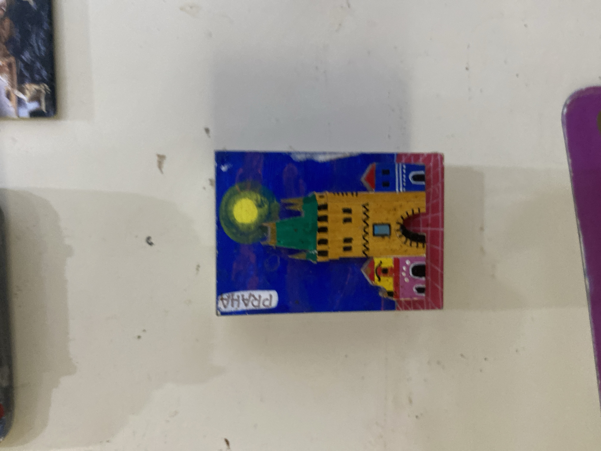A hand-painted refrigerator magnet depicts a simplified depiction of a tower and surrounding buildings against a blue sky with a yellow sun. The word "PRAGA" is written vertically on the left side of the magnet. The magnet is affixed to a white, textured surface that appears to be a refrigerator door. The image is blurry and taken from a slight angle. There is no indication of the city of Volzhsk, Russia.