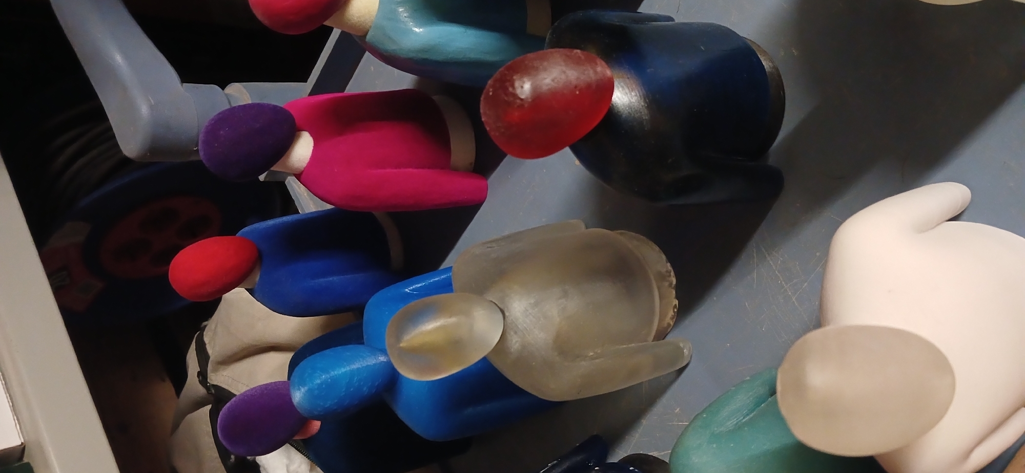 The image shows a collection of colorful, stylized human figures, possibly figurines or sculptures, arranged on a gray surface. The figures feature spherical heads in colors like red, purple, and clear, attached to bodies in shades of pink, blue, and turquoise. The figures appear to be lined up or stacked together. The photo is taken from an elevated perspective, looking down on the figures. The scene includes a metallic object on the left, possibly part of a structure or machinery, as well as hints of fabric and other objects in the background. The location is not provided by the image.
