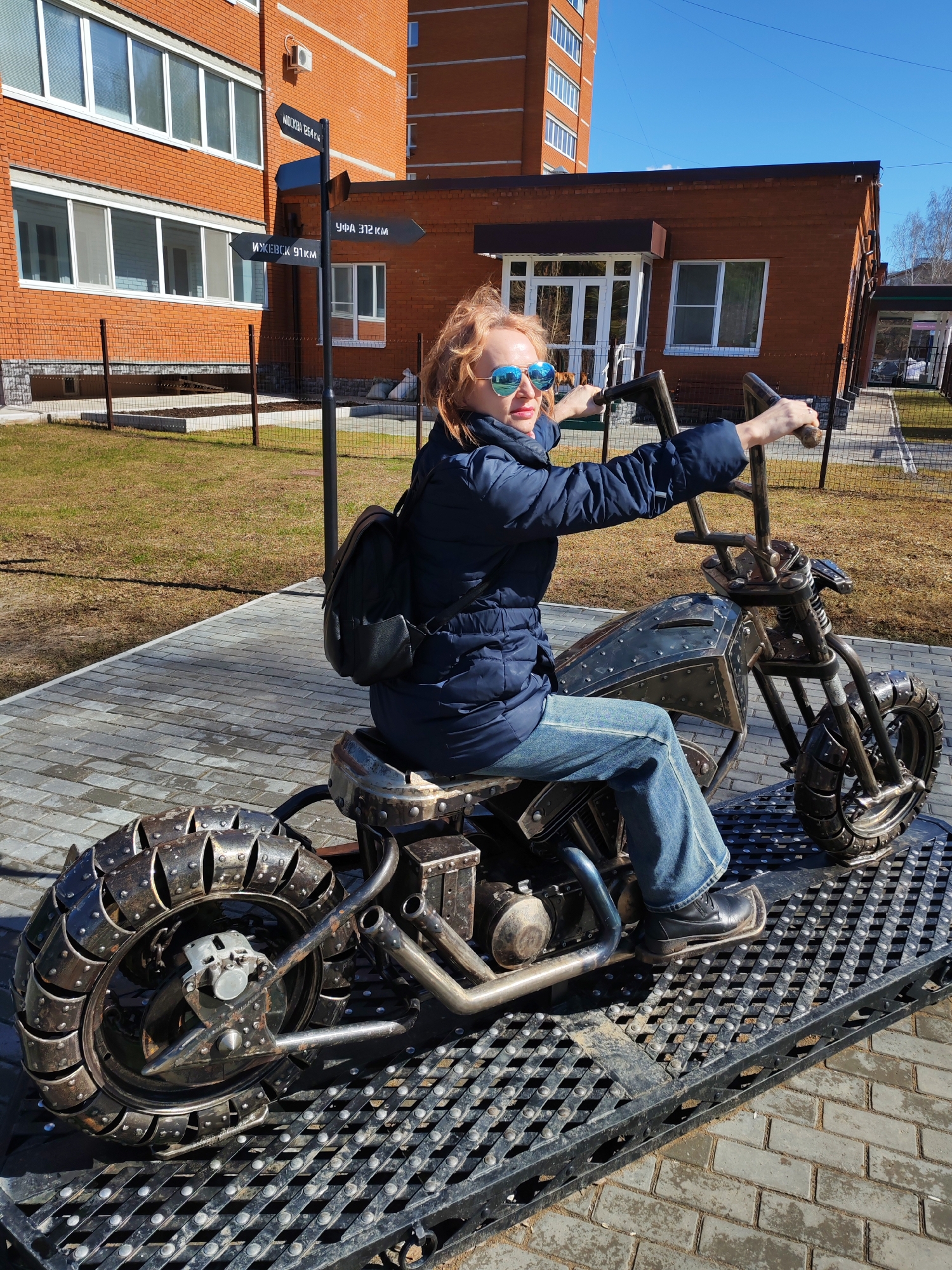 A woman wearing blue mirrored sunglasses, a blue jacket, jeans, and black shoes is sitting on a metal motorcycle sculpture in the city of Chaikovsky, Russia. She has a black backpack on her back and is holding the handlebars of the motorcycle. The motorcycle is made of metal and has a unique design, including the wheels. The sculpture is placed on a metal platform. In the background, there are brick buildings, a directional sign indicating distances to Moscow, Ufa, and Izhevsk, and patches of grass. The sky is clear and blue. The scene appears to be a sunny day in a public area.
