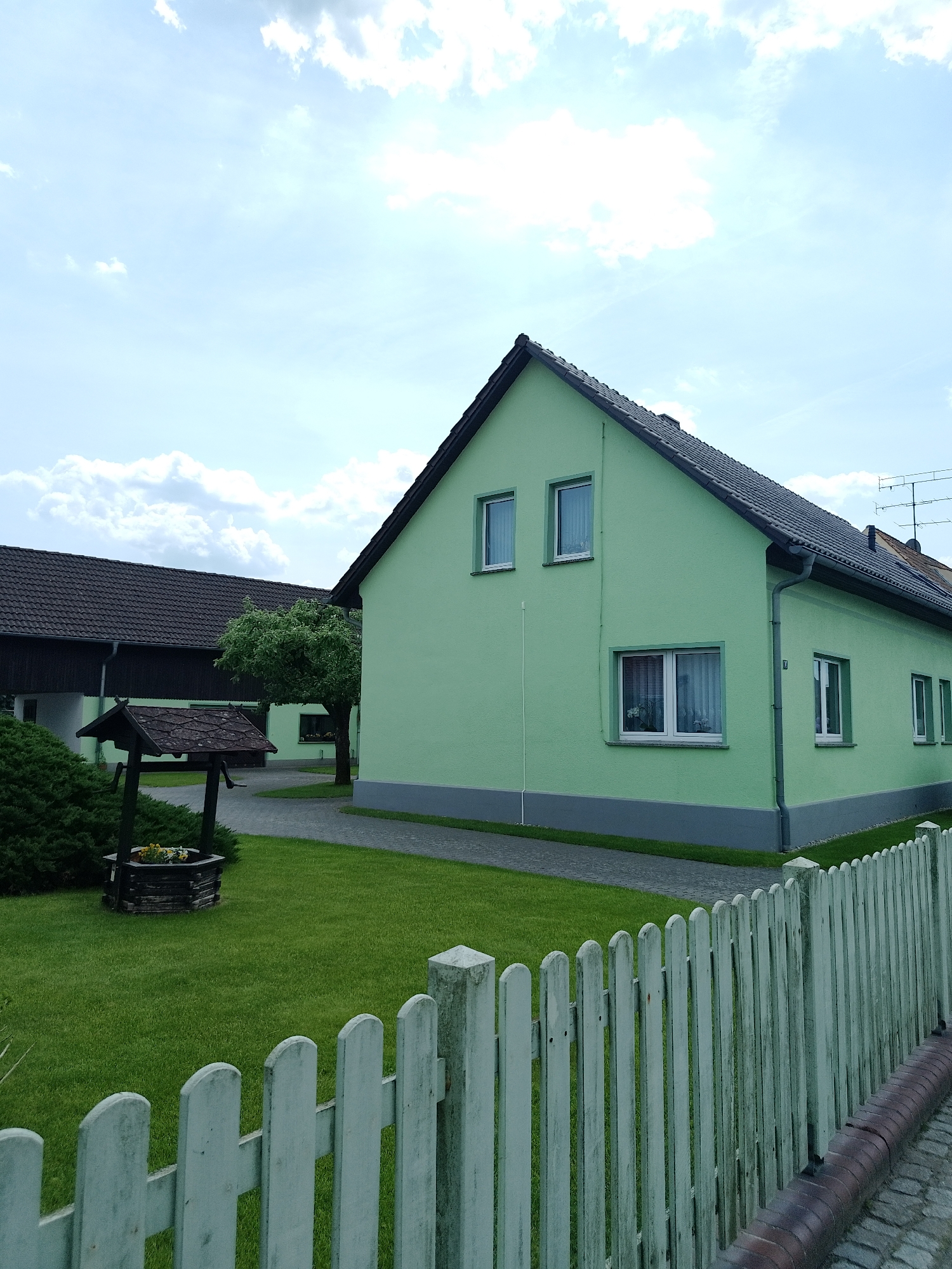 The image shows a residential scene in an unknown location, likely a small town or village. The foreground is dominated by a weathered white picket fence running along a brick-edged path. Behind the fence is a well-maintained lawn. To the left of the lawn is an ornate wooden well, surrounded by a green bush. Behind the well is a two-story green house with white window frames and a gray trim along the base. To the left of the green house is a large, dark-roofed building. A tree sits between the green house and the darker building. The sky is blue with scattered white clouds.