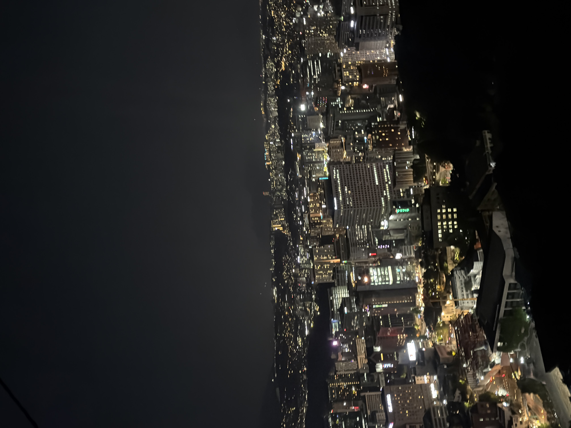 The image depicts a nighttime cityscape, likely in Seoul, South Korea, viewed from an elevated position. The left side of the image shows a dark, open area, possibly the Han River or a large park. Moving right, the cityscape unfolds with densely packed buildings illuminated by numerous lights, forming a grid-like pattern. The buildings vary in height and size, suggesting a mixed-use urban environment. The lighting indicates both residential and commercial activities. The foreground is dark, obscuring details, but hints at nearby structures. Overall, the image captures the scale and intensity of urban life at night in Seoul.