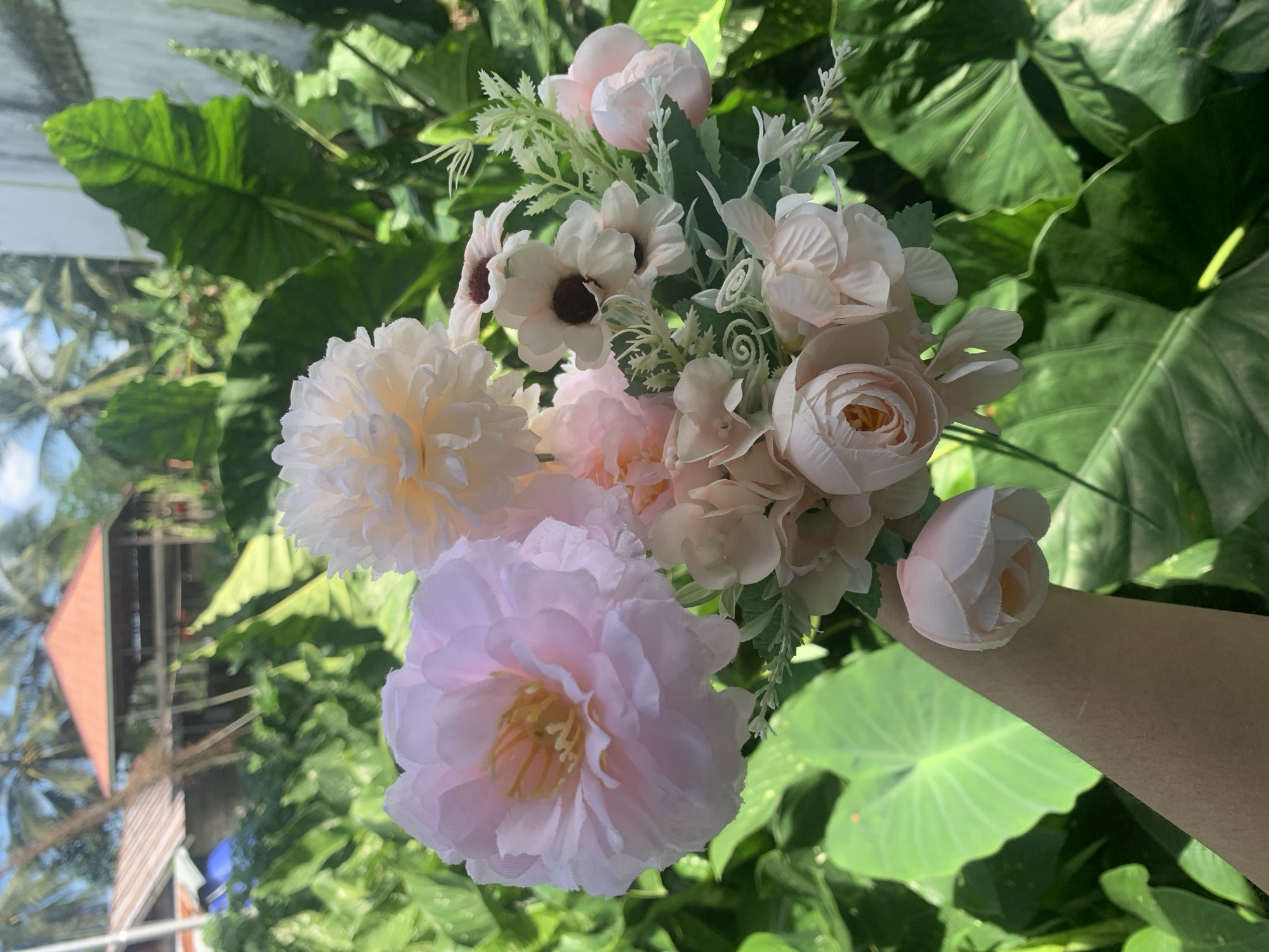 A bouquet of artificial pink and white flowers is held in a person's arm, in front of a lush green outdoor environment. In the background, out of focus, there are palm trees and two buildings with orange roofs. The scene appears to be set in a tropical location, potentially Kuala Pembuang, Indonesia.