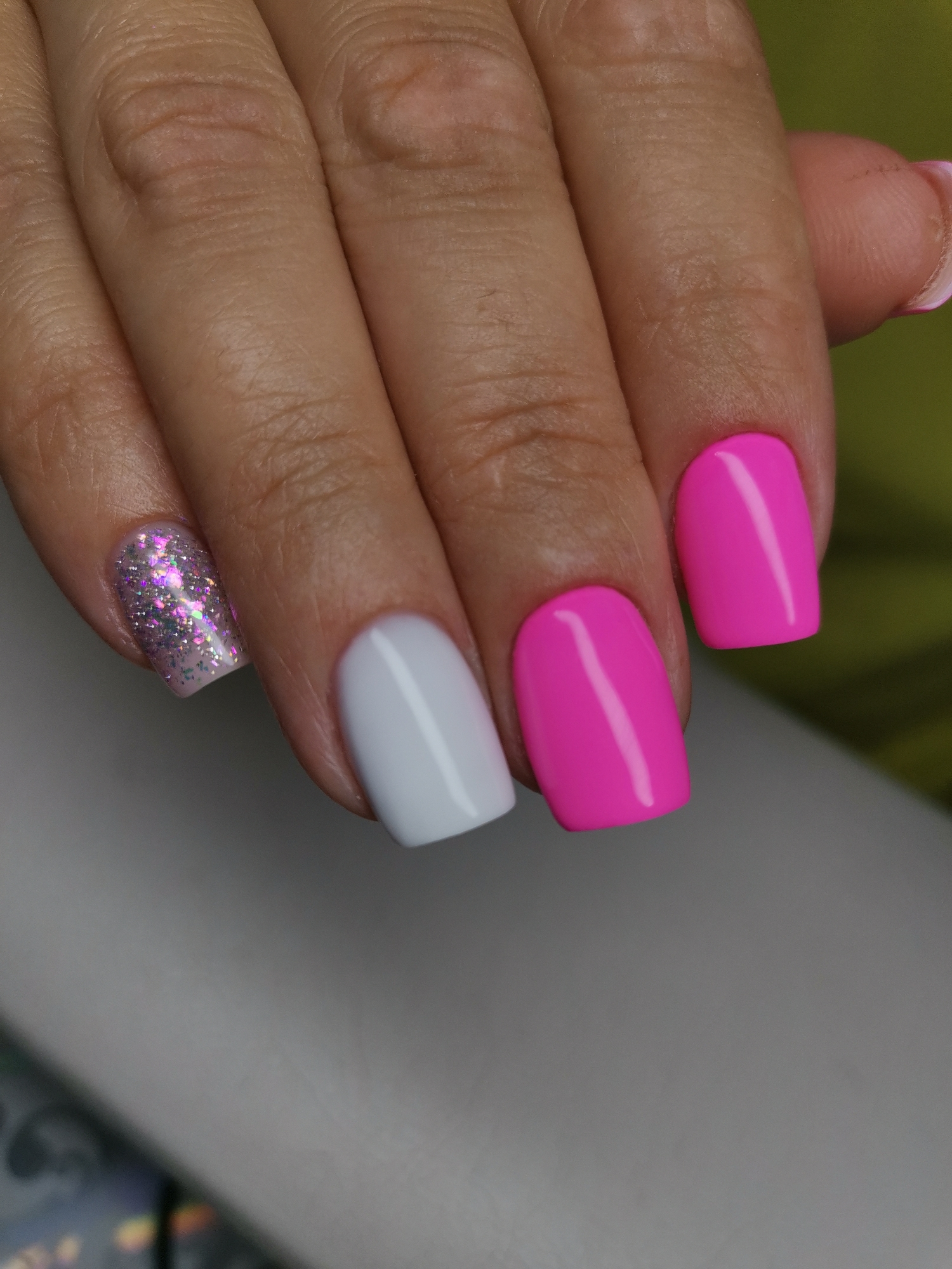 The image shows a close-up of a person's hand with manicured nails. The nails are painted in a variety of colors and finishes. The index finger has a white polish, the middle finger has a bright pink polish, and the ring finger has a glittery pink polish. The thumb and pinky finger are mostly out of frame, but the pinky finger appears to have a pink polish as well. The background is a blurred green surface on the right and a light gray surface on the bottom. The lighting suggests an indoor setting. There is no indication of the city of Kizel, Russia, or any specific environmental details that would place it there.