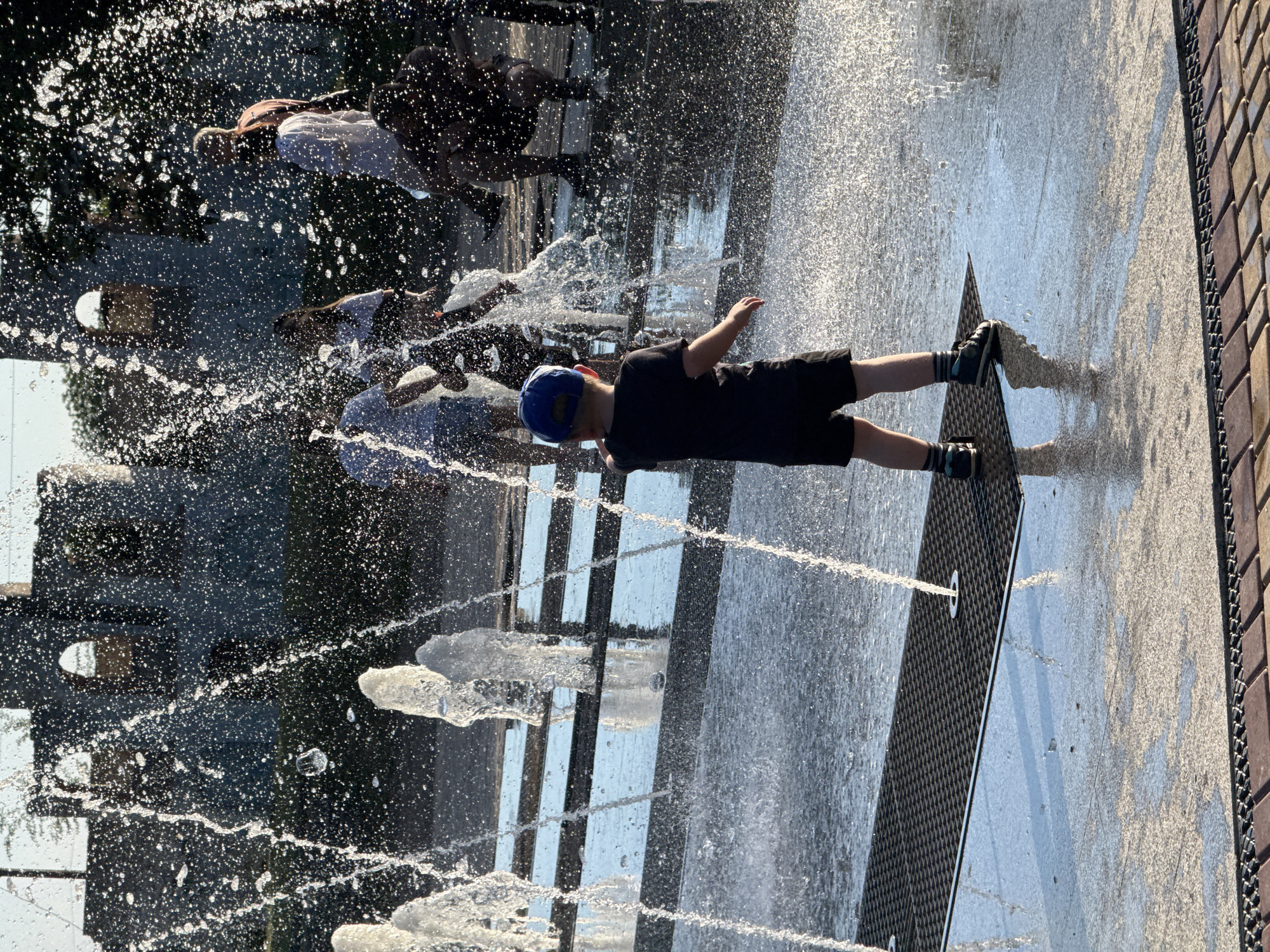 In the city of Unknown, Unknown, a young boy wearing a blue cap, black t-shirt, and black shorts stands on a grate in a splash pad. Water sprays around him and other people in the background. The background features a stone structure with arched openings and trees. The ground is wet, reflecting the sunlight.