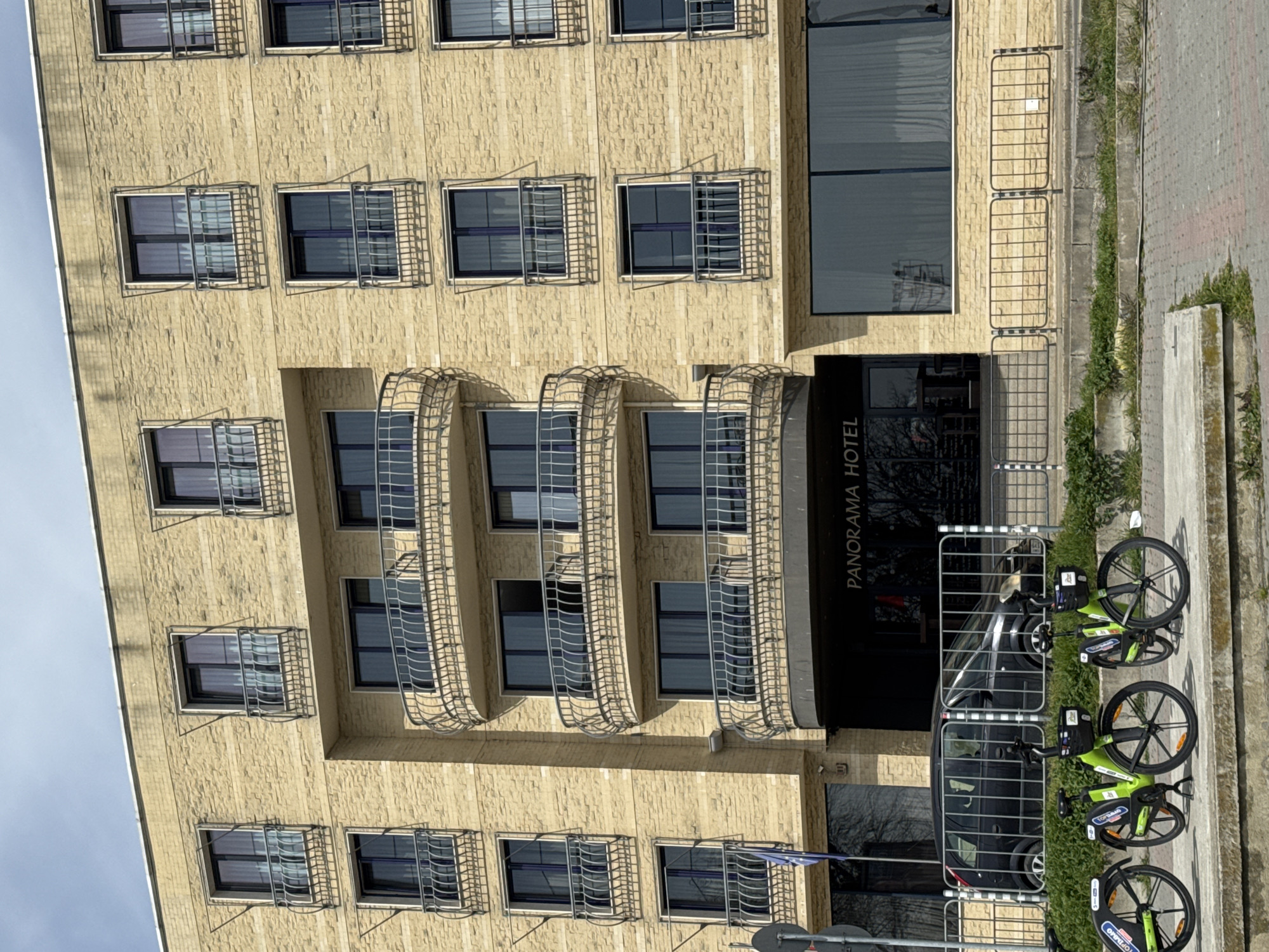 The image shows the Panorama Hotel in Varna, Bulgaria. The hotel is a light-colored brick building with multiple stories of windows and decorative metalwork. The hotel entrance is under a dark awning with the words "PANORAMA HOTEL" in white lettering. There are several green and black rental bicycles parked along the sidewalk in front of the hotel. A temporary metal barricade is also present. There are no people visible in the image. The sky is visible in the upper left corner of the image.