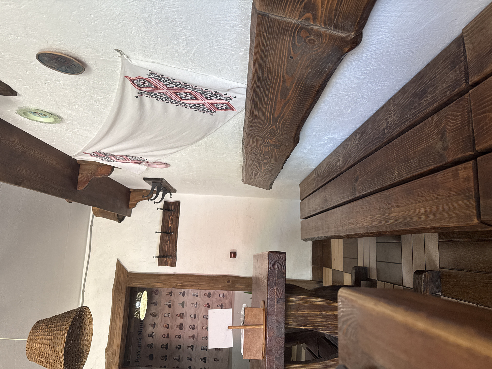 The image shows a rustic interior, likely a restaurant or cafe, with wooden furniture and white walls. In the foreground, a heavy wooden table and bench are prominent, suggesting a traditional or folk-inspired theme. A woven lampshade hangs from the ceiling to the left. On the wall, a white cloth with red and black embroidered patterns is displayed, along with a framed collection of portraits. There are no people visible in the image. The floor is tiled in a checkered pattern of brown and beige. The environment appears well-lit, with natural light coming from an unseen source. There is no specific information within the image that places it in Noviy, Russia.