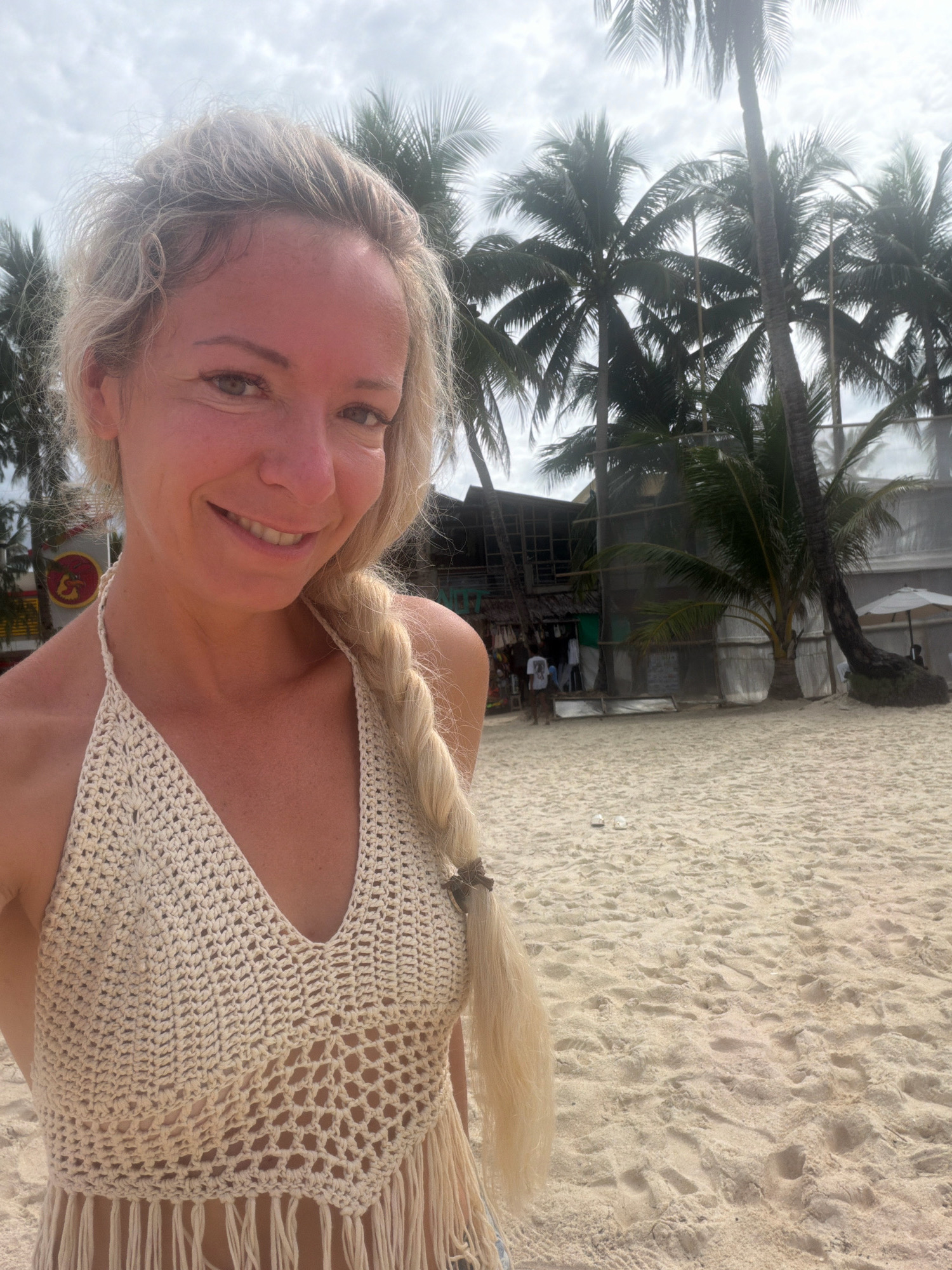 A blonde woman with a braid smiles at the camera, her upper body filling the left half of the frame. She is on a sandy beach, with palm trees and a building visible in the background. The woman wears a beige crocheted halter top with fringe. The building in the background appears to be a small shop or restaurant with a thatched roof. There are other people and a white umbrella visible in the distance. The sky is cloudy. This is a common scene on Boracay Island, in the city of Malay, Philippines, known for its white sand beaches and palm trees.