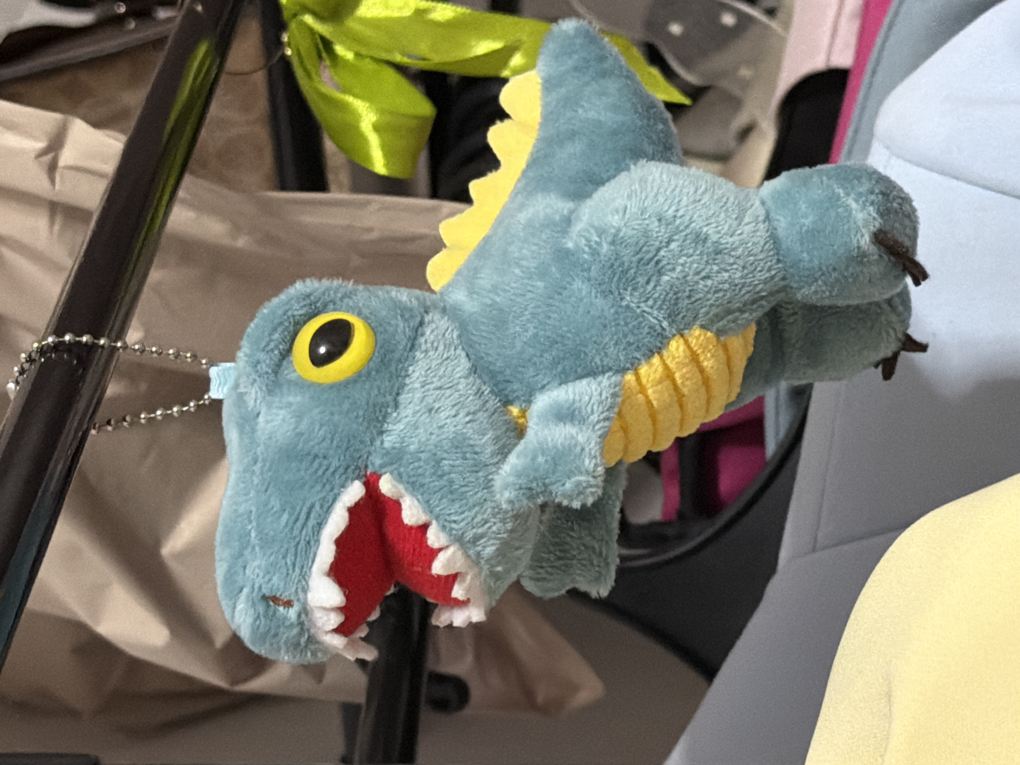 A close-up, eye-level shot shows a teal-colored plush toy dinosaur with yellow accents and red mouth details hanging from a metal bar. The dinosaur is suspended by a silver beaded chain. The background is out of focus, but appears to be a domestic or retail setting with fabric draped and a bright green ribbon tied to the bar. The lighting is bright, casting some shadows. The image was taken in Moscow, Russia.