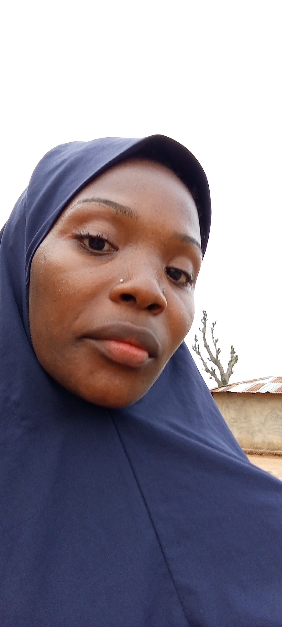 A close-up shot shows a woman wearing a dark blue hijab. She has dark skin and a nose piercing. Behind her, there is a building with a corrugated metal roof and a bare tree. The picture was taken in the city of Keffi, Nigeria.