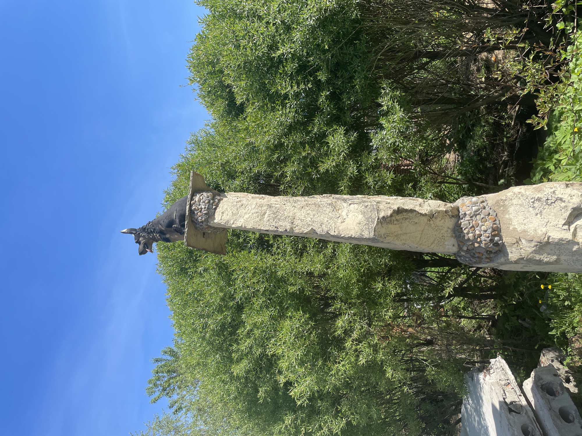 A vertical shot depicts a sculpted gargoyle-like animal figure perched atop a weathered stone pillar. The pillar, seemingly constructed of roughly hewn stone and pebbles, stands against a backdrop of lush, green foliage and a clear, blue sky. Parts of broken concrete blocks can be seen to the right, at the base of the pillar. Based on online information, this might be located in Perm, Russia.
