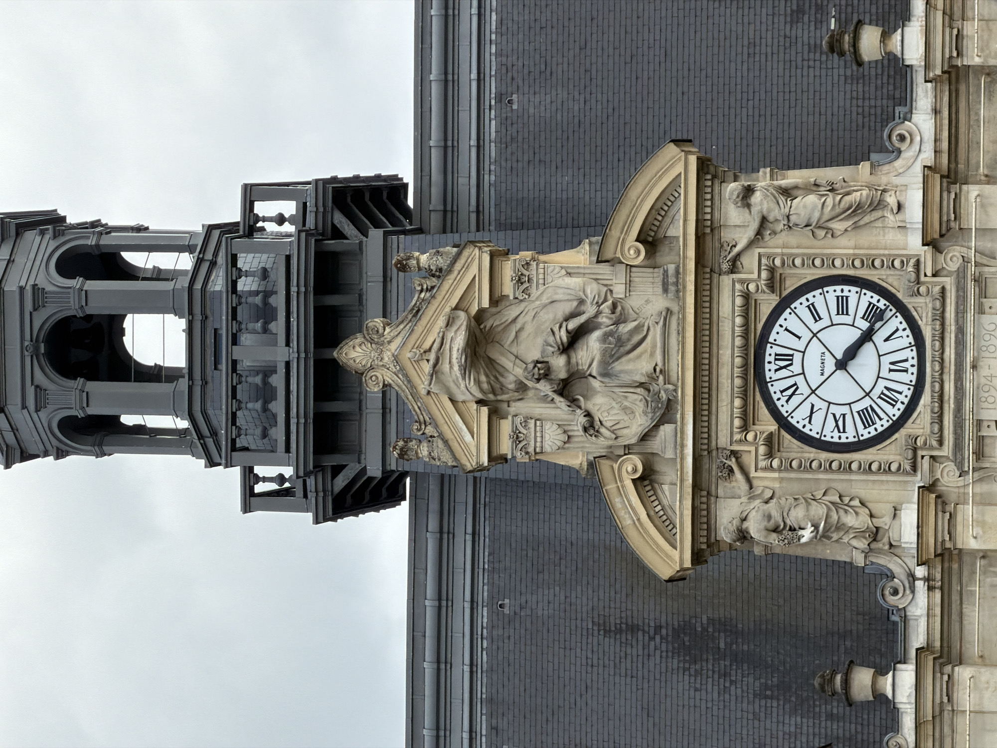 This media file displays a close-up, vertical view of the facade of a building, likely a public building or clock tower, in Ivry-sur-Seine, France. The primary subject is an ornate clock face with Roman numerals and black hands, indicating the time is approximately 3:50. Flanking the clock are detailed stone sculptures of classical figures. One figure, positioned above the clock, reclines while reaching out. Another figure, below the clock, also reclines and holds what appears to be grapes. The architecture features elaborate stone carvings, arches, and decorative moldings, rendered in a light-colored stone, contrasted with a dark, brick-patterned surface on the building's upper sections. To the left of the clock and sculptures is a section of dark, tiered architectural elements, possibly a tower or spire, with open arched sections. The sky is overcast and a light gray, suggesting a cloudy day. Visible text on the clock face reads "MAGNETTA" and "1894-1896" is inscribed vertically on the right side of the building's facade.