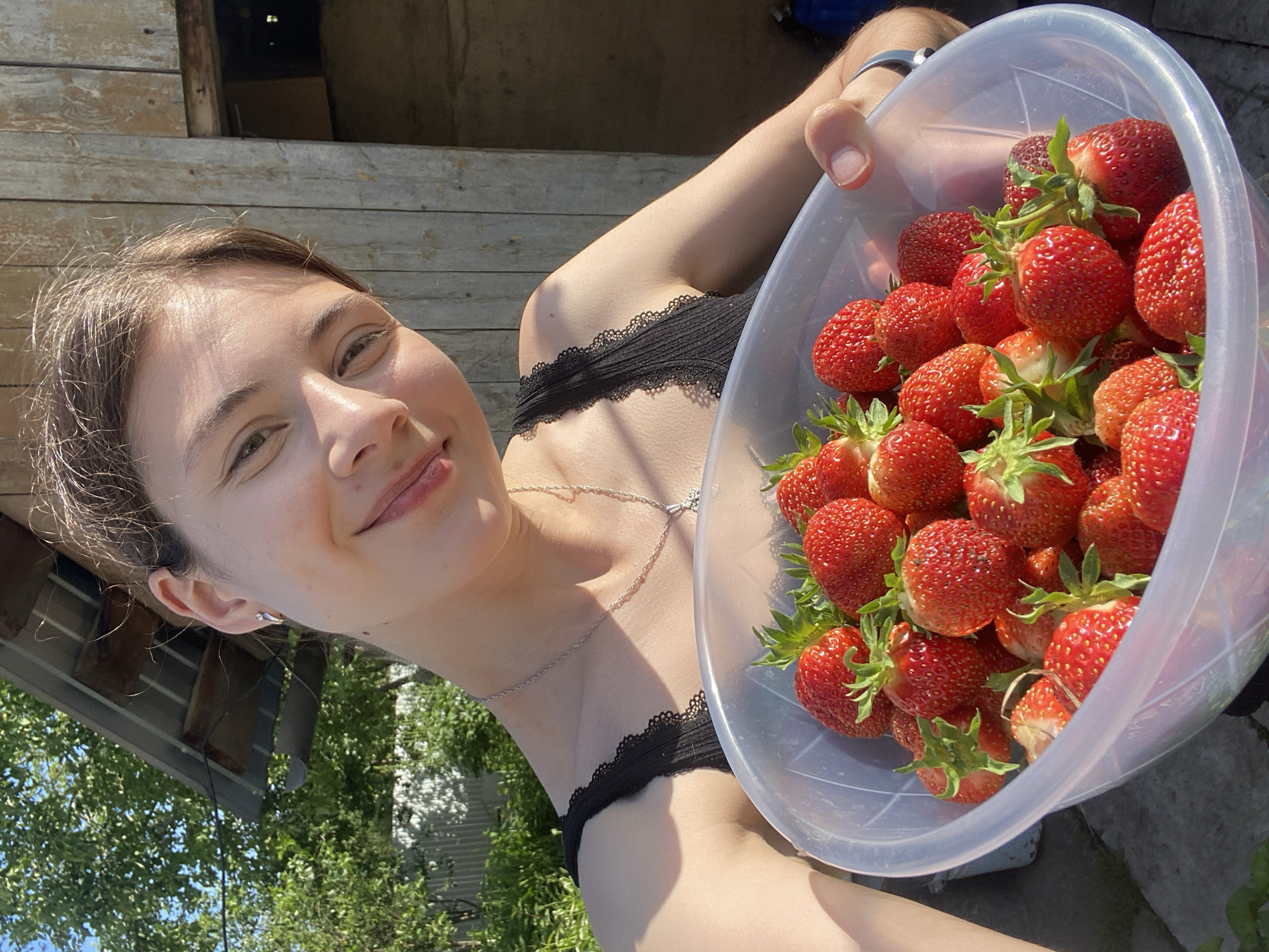 A young woman with light brown hair, wearing a black lace-trimmed tank top and a silver necklace, holds a large clear bowl overflowing with ripe strawberries. She is outdoors, with a rustic wooden structure and green foliage in the background, suggesting a rural or suburban setting. The lighting indicates it is daytime, likely sunny. The woman is smiling and looking towards the camera as she presents the bowl of strawberries. The location is Votkinsk, Russia.