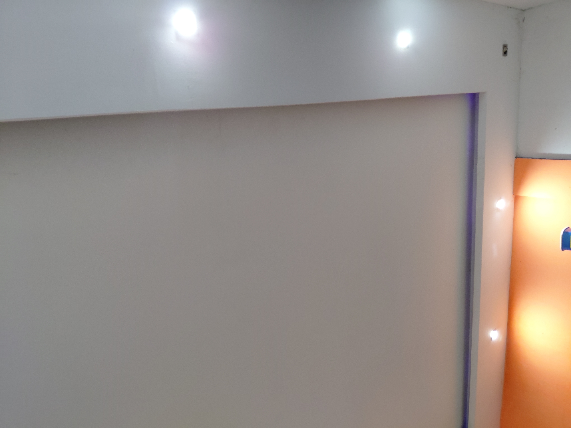The image shows a section of an interior wall with a white top border and a large, light gray rectangular area framed with a thin, purplish border on the right. The wall abuts an orange section with two small spotlights. Three spotlights are visible on the top border. A corner is visible at the top right. A small rectangular object is visible on the top right corner of the image.
There is no indication of the location being in the city of Santa Ana, Venezuela, nor any people, actions, interactions, or other objects visible in the image.