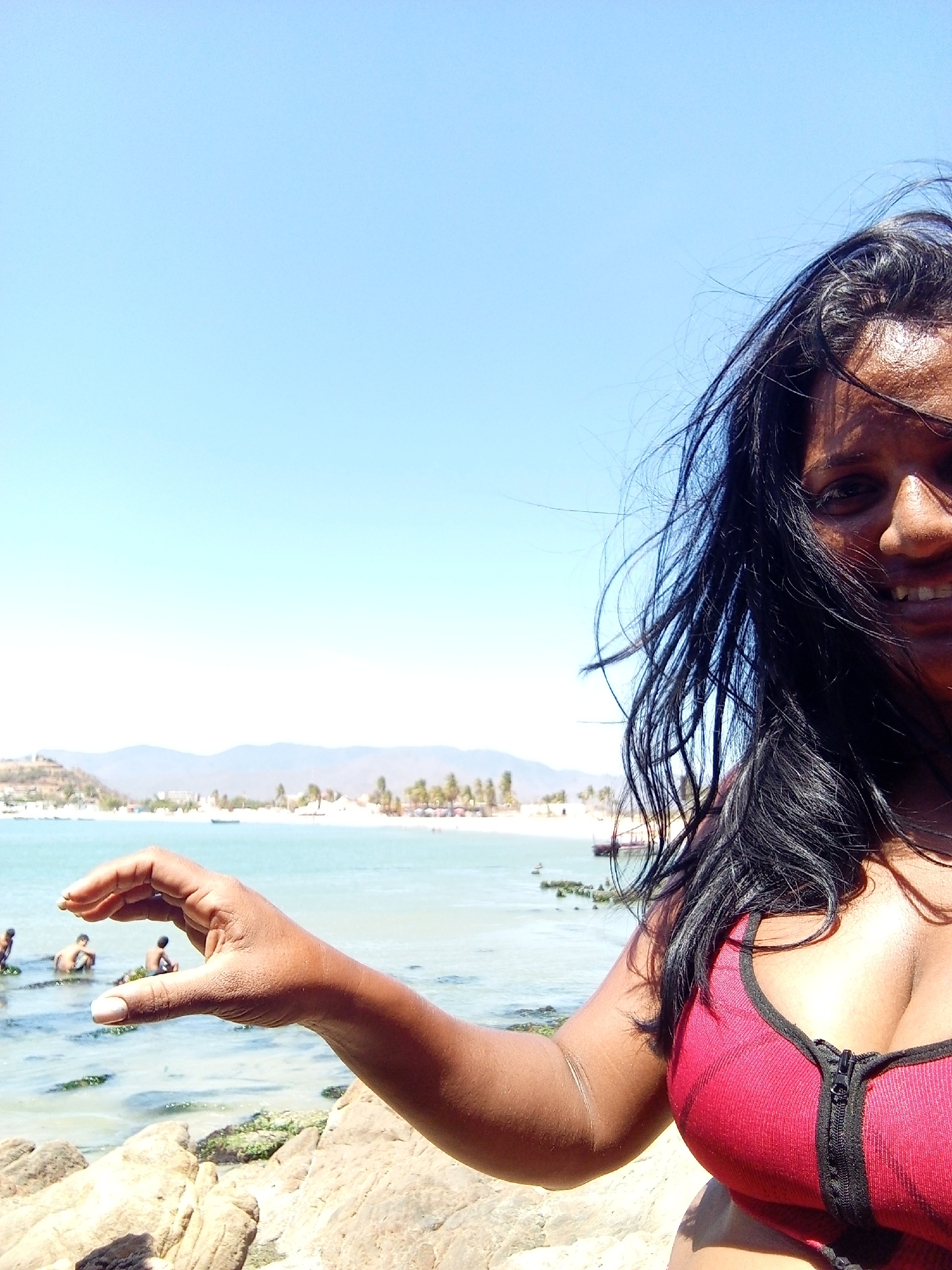A selfie taken on a sunny day at a beach in Juan Griego, Venezuela. The photographer, a smiling woman with long black hair, is wearing a red top. Her right arm is extended, her hand making a circle, as if framing the beach. In the background, several people are standing in the shallow water. The beach is sandy with some rocky areas and some green algae is visible. In the far distance, the town of Juan Griego is visible against a backdrop of mountains and a clear, blue sky.