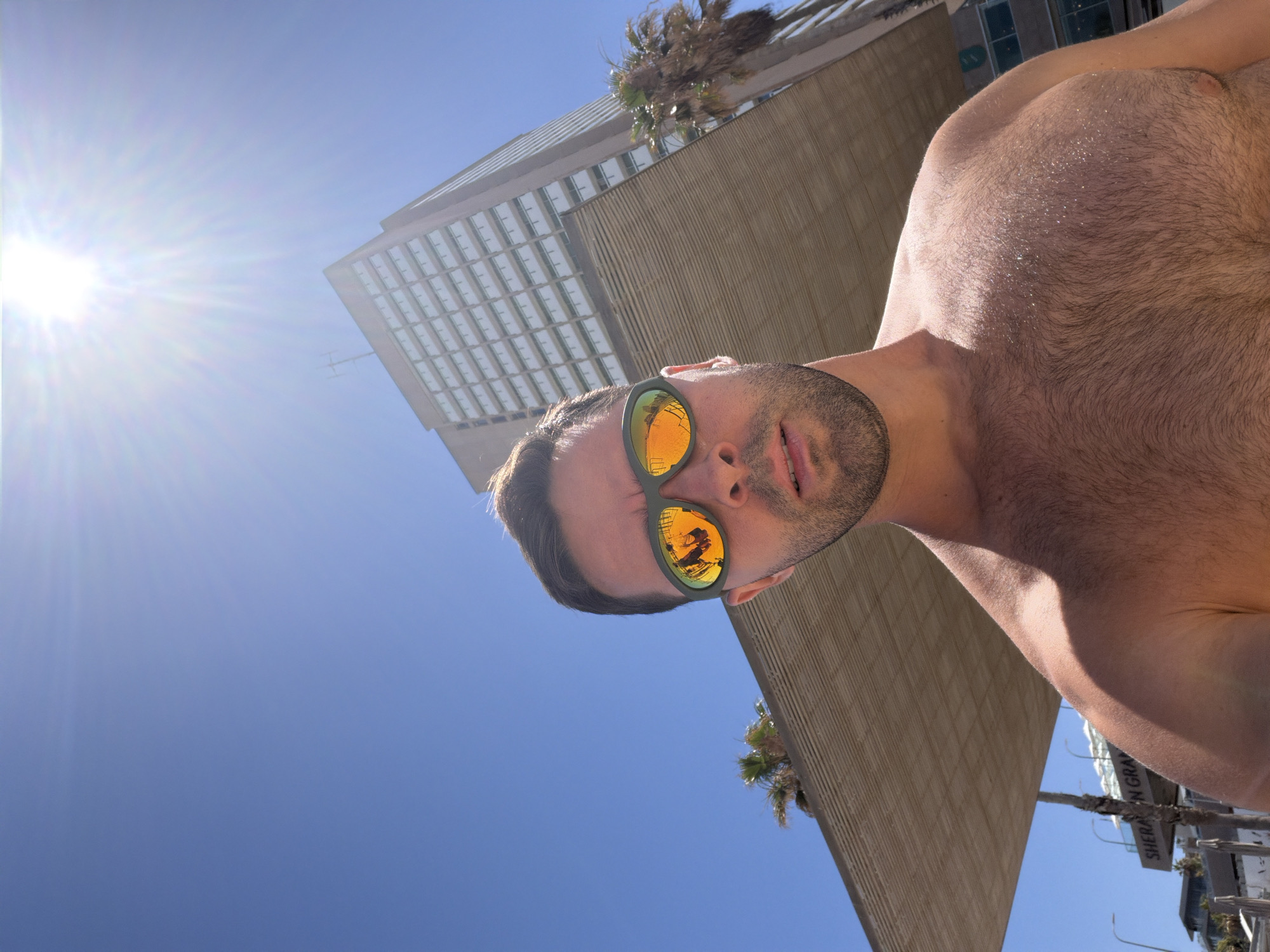 The image is a bright, low-angle selfie taken on a sunny day, with the frame rotated 90 degrees clockwise. It features a shirtless man looking directly at the camera. His face, angled slightly upwards, is positioned on the left side of the frame. He has short dark hair and stubble, and is wearing green-framed sunglasses with reflective orange lenses. His upper torso, including his chest and shoulder, is visible on the right, showing dark body hair that glistens in the strong sunlight.
The background is dominated by a clear, bright blue sky, with a dazzling sunburst emanating from the top-left corner, indicating strong daylight. Behind the man, an urban landscape unfolds. A tall, modern building with a grid of rectangular windows or panels is prominently featured. Adjacent to it, another section of a building displays a textured, ribbed, light brown facade. Several palm trees are scattered throughout the background, hinting at a warm climate. In the bottom-right corner, a partial sign with the text "SHERA" and "GRA" is visible, strongly suggesting a "Sheraton Grand" hotel.
The scene depicts a relaxed moment in a vibrant, sunny city environment, characteristic of Tel-Aviv, Israel, particularly given the presence of a Sheraton Grand hotel in the city. The time of day appears to be midday or early afternoon due to the intense sunlight.