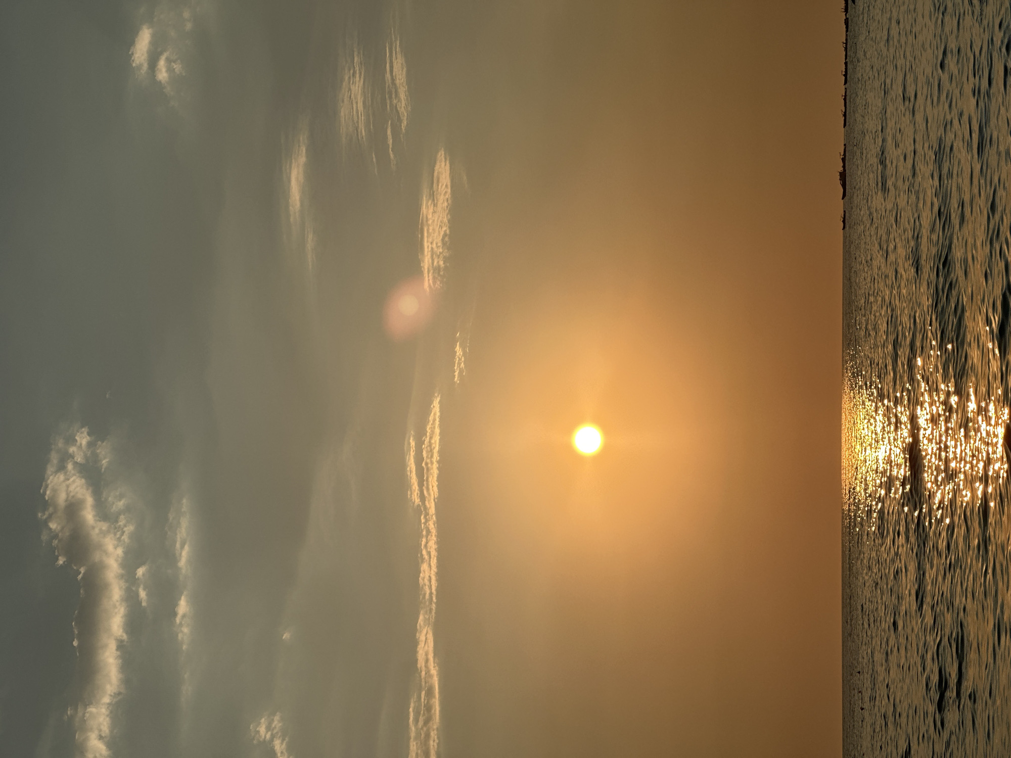 An outdoor, vertical, full shot captures the sun setting over a calm body of water. The sun is a bright, orange orb, positioned slightly left of the center of the frame, radiating a golden glare. The sky is a hazy, warm orange, with some scattered clouds appearing as streaks of white and grey. The water in the foreground is dark and slightly rippled, reflecting the sun's light in a shimmering path across its surface. On the far right horizon, a line of small boats is visible, silhouetted against the sunset. The overall atmosphere is serene and tranquil.