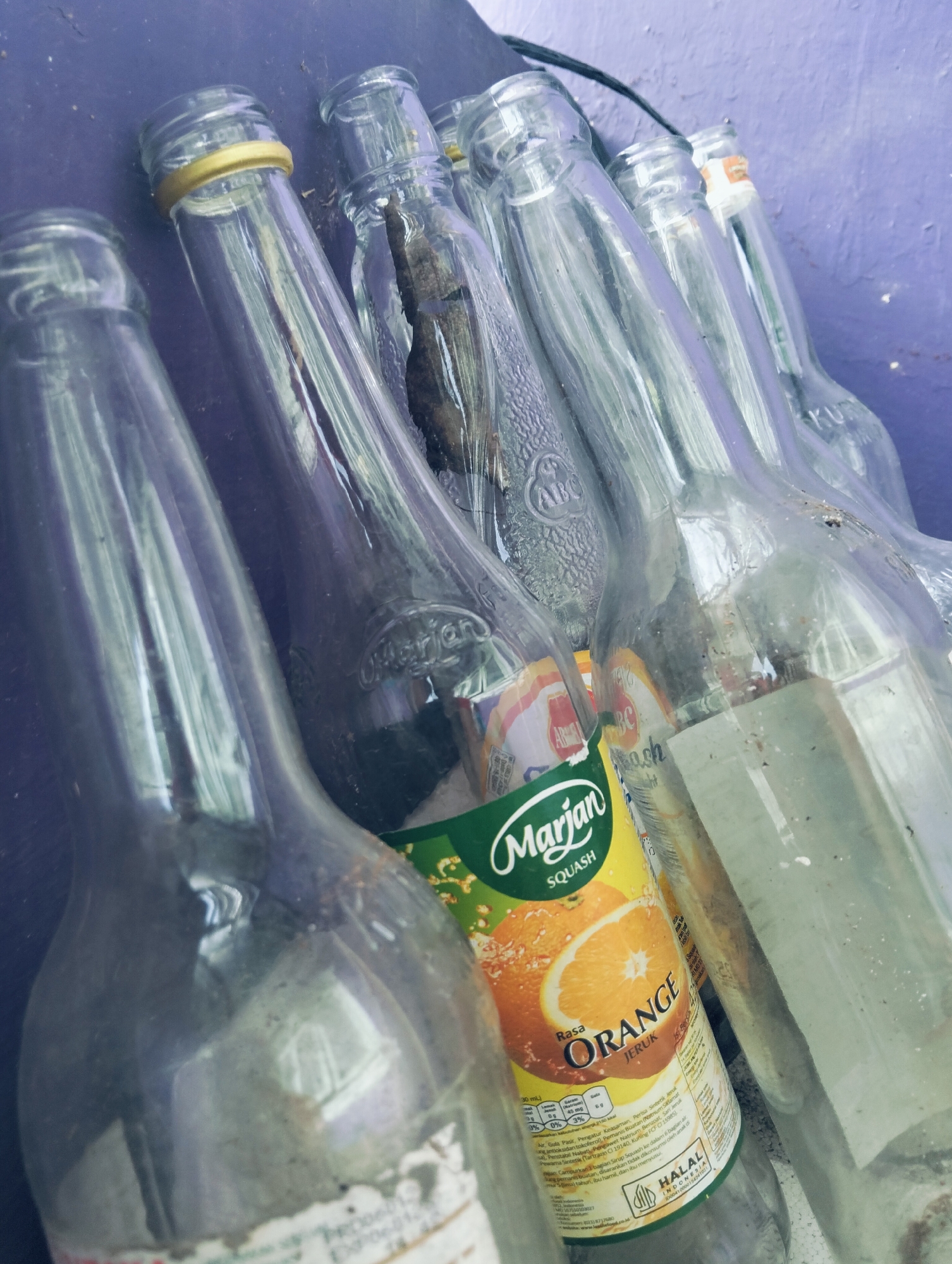 A group of clear glass bottles, some with labels, are arranged in a row on a dark surface, possibly a table or floor, against a textured purple wall. The bottles are mostly empty, with some residue visible inside some of them. One bottle has a prominent label for "Marjan Squash Orange." The lighting appears to be overhead, casting reflections on the glass. The scene is indoors or in a covered outdoor area, given the wall and the arrangement of objects. There are no people or animals present. The environment appears somewhat dusty or aged. The overall impression is one of discarded or stored beverage containers.