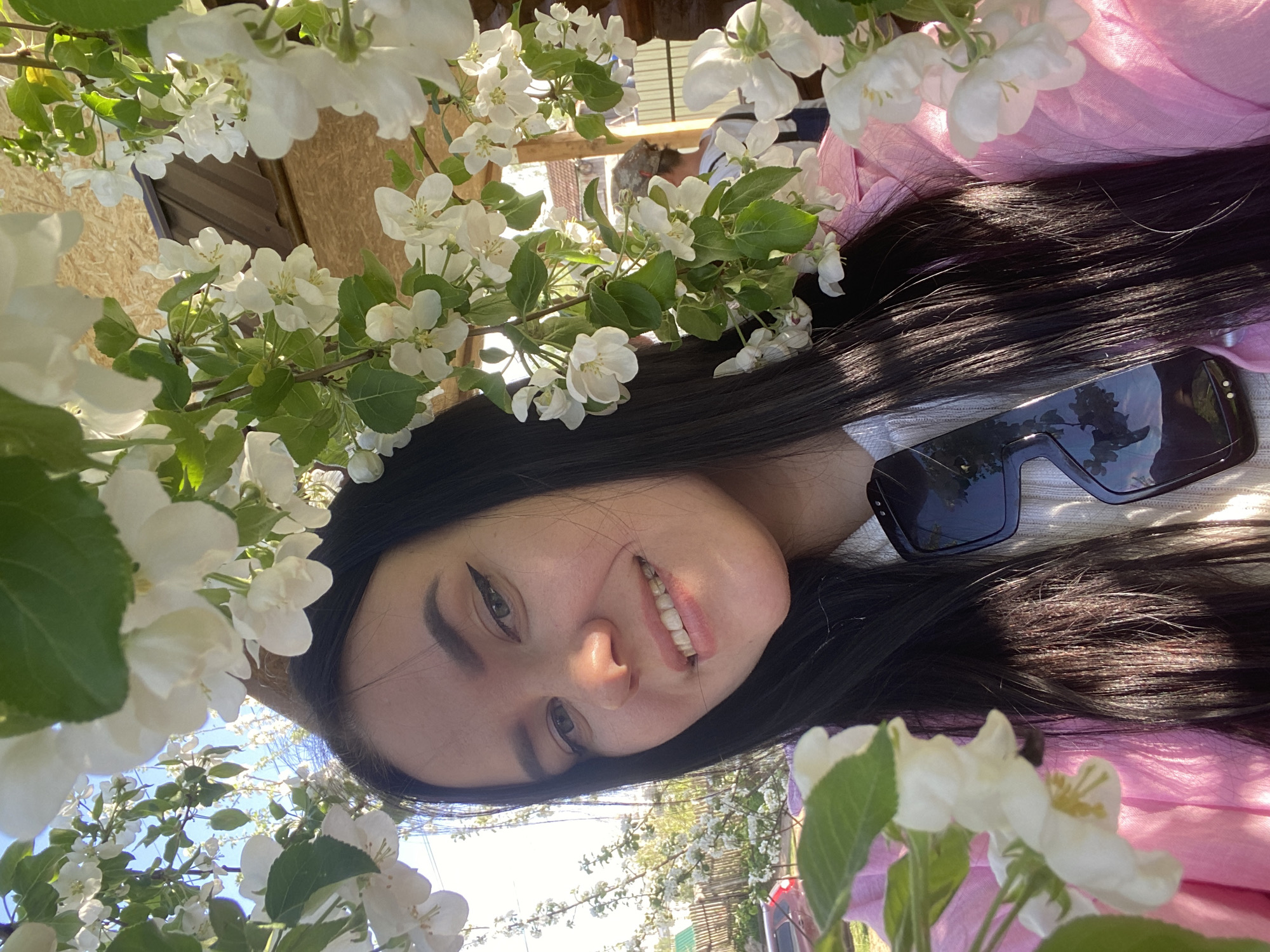A young woman with dark hair and sunglasses is the primary subject of the image, smiling slightly at the camera. She is surrounded by white blossoms, possibly from an apple tree, with green leaves visible. The background shows hints of a wooden structure and greenery. The presence of a pink garment draped around her suggests a casual, outdoor setting, possibly a park or garden in Chaikovsky, Russia. There are no obvious indications in the image to confirm it is Chaikovsky Russia.