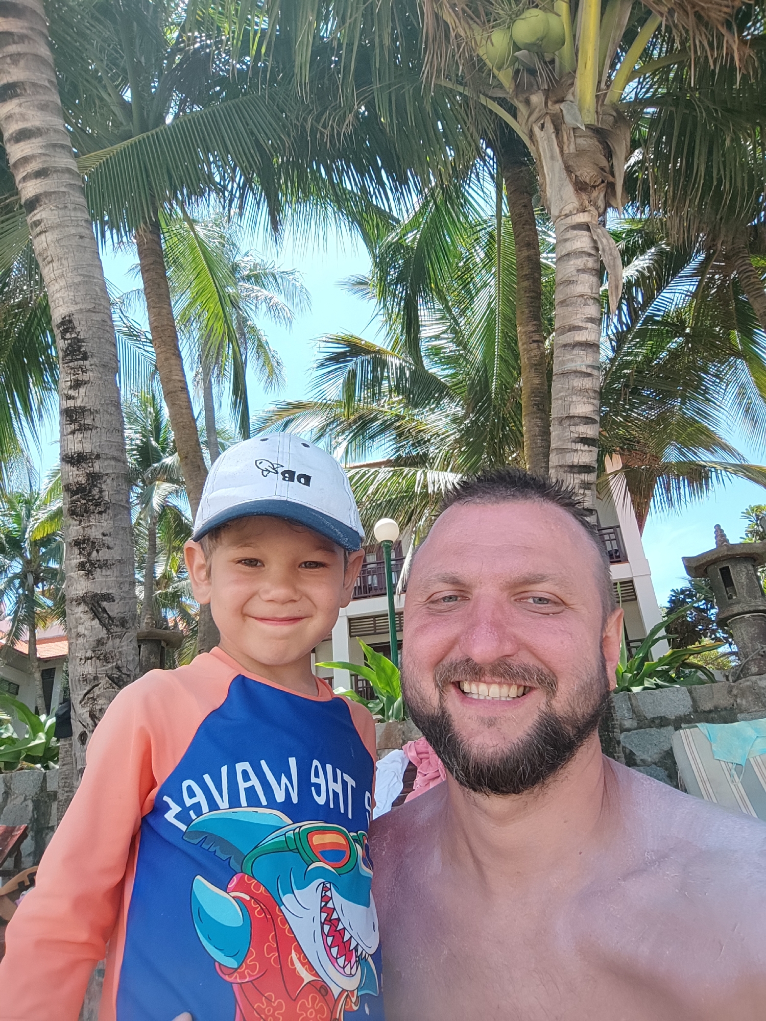 A father and son are captured in a selfie outdoors. The son, wearing a white baseball cap and an orange and blue rash guard with a shark graphic, stands to the left. The father, with a beard and a tan, is on the right. They are surrounded by palm trees and a building in the background, suggesting a tropical resort setting. The scene appears to be in Phu Thuy Ward, Vietnam.