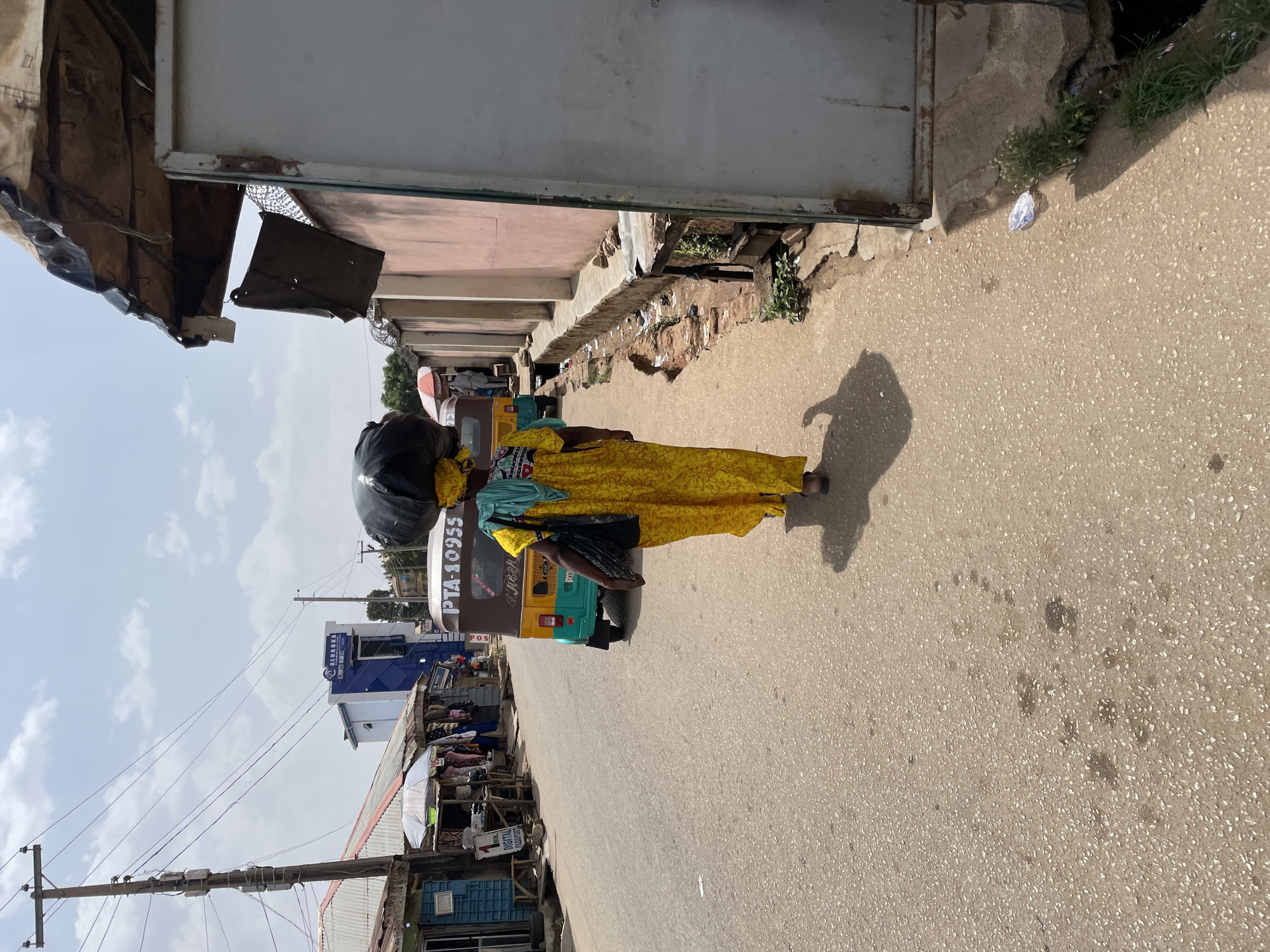 A woman in a yellow patterned dress walks along a paved road in Jos, Nigeria. She carries a large, black object on her head. To her left, there is a drainage ditch and a line of shops with varying structures, including a building with a blue and white sign that says "Albarka". An auto rickshaw, with the license plate PTA-10955, is beside her. Overhead, there are electrical wires connected to wooden poles. The sky is pale with clouds.